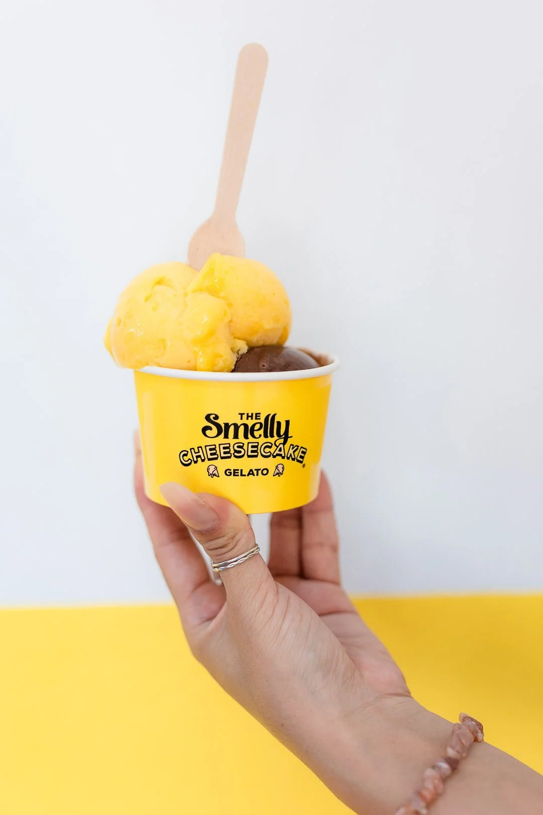 Person holding a yellow cup with the text 'The Smelly Cheesecake Gelato,' filled with yellow and brown gelato and a wooden spoon, against a split white and yellow background.