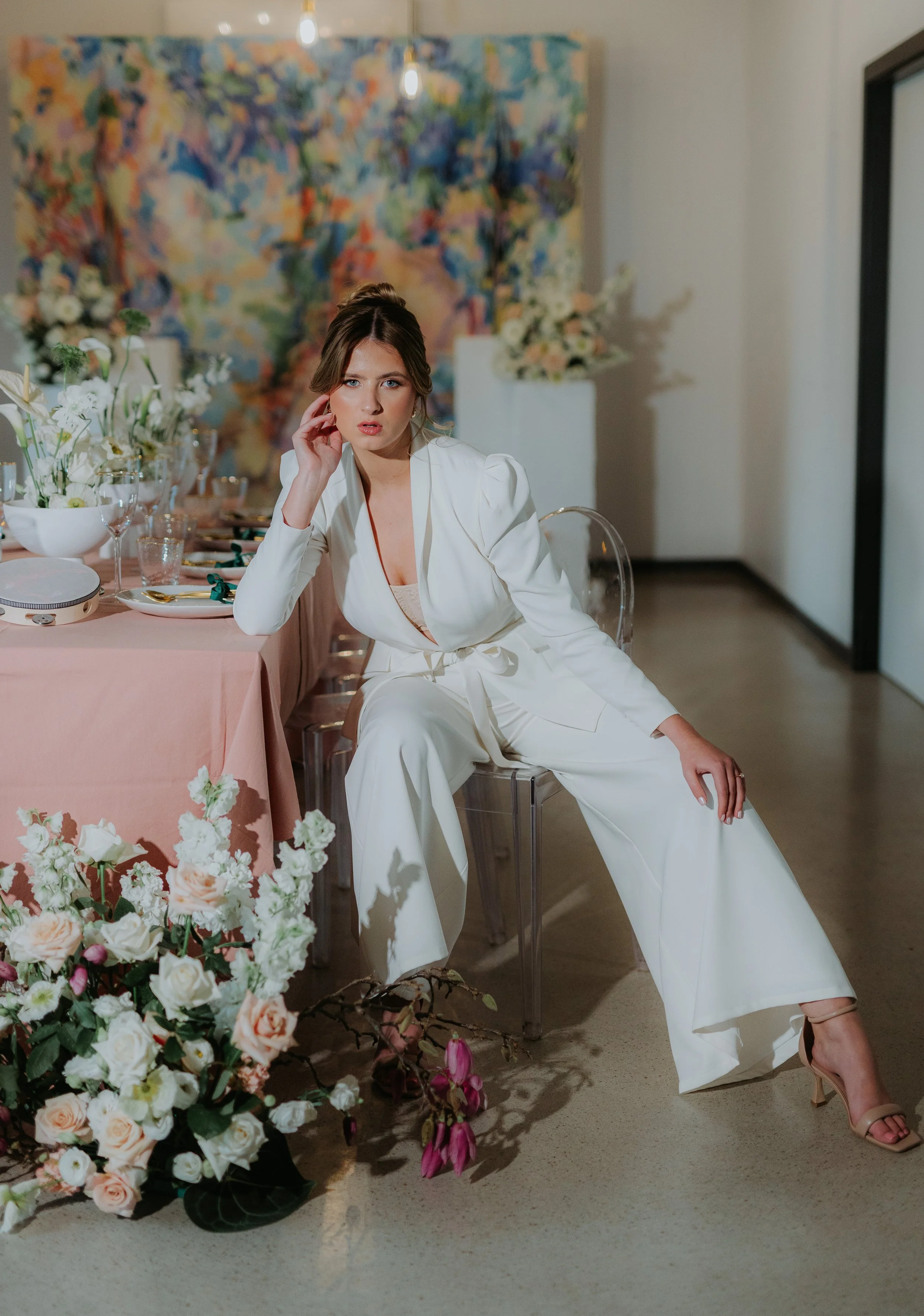 A woman in a white suit sitting at a decorated dining table with flowers, in an art gallery setting.