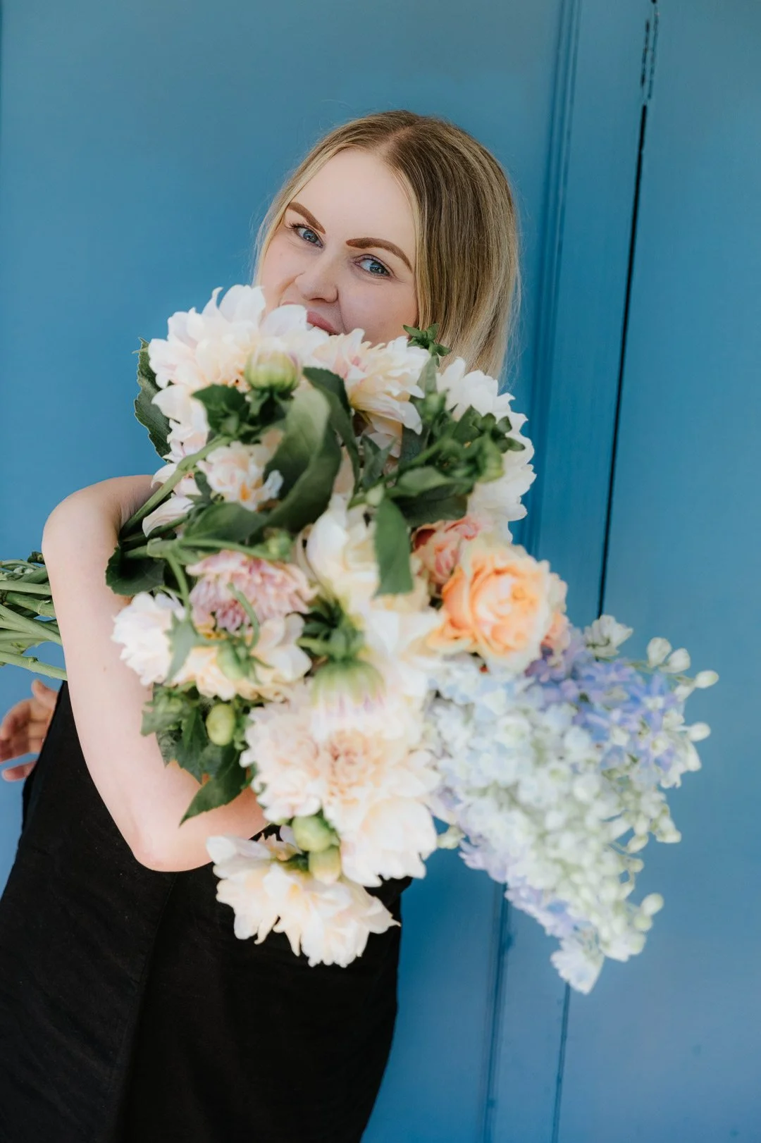 A woman with blonde hair and blue eyes holding a large bouquet of pastel-colored flowers in front of a blue background.