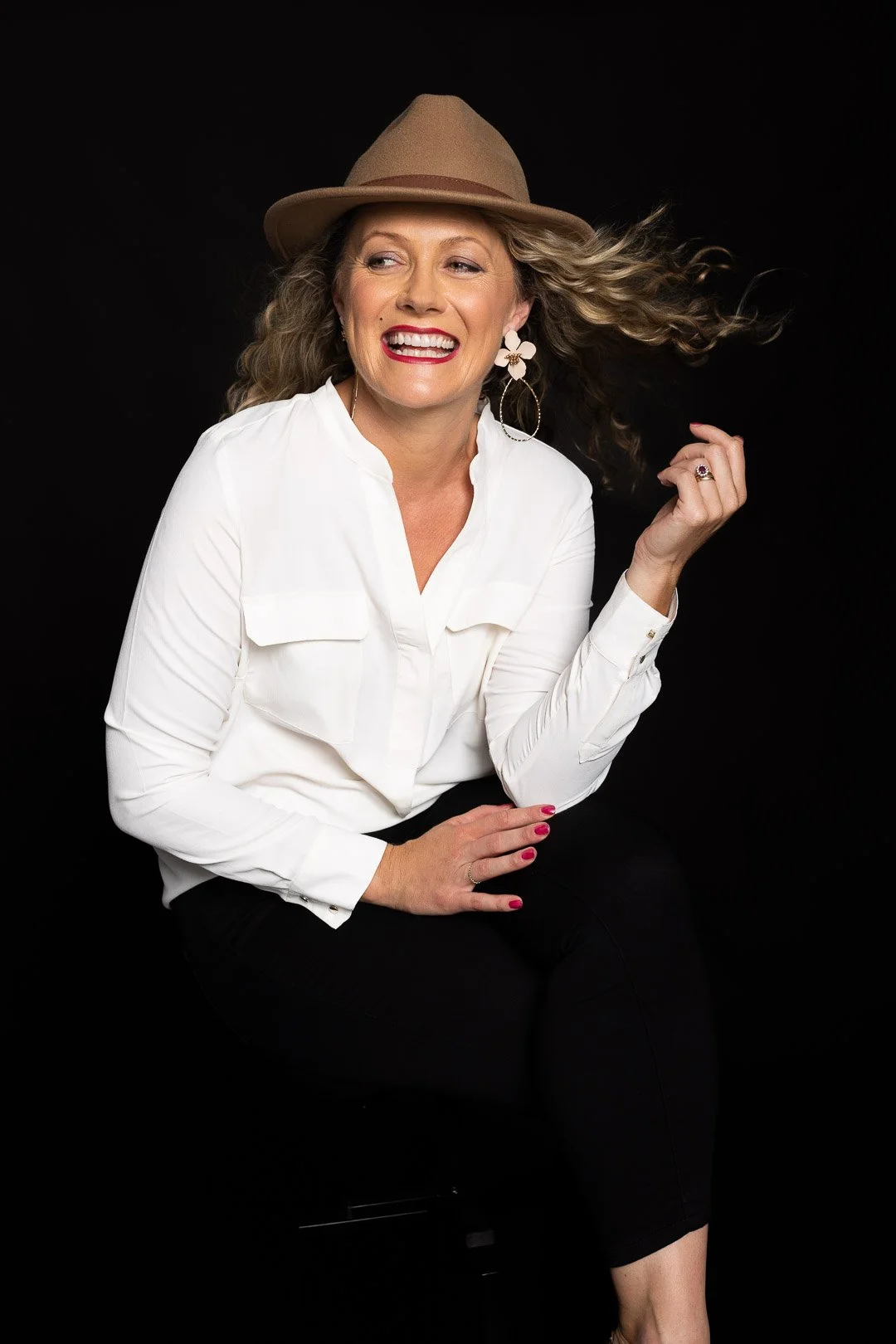A woman with curly hair wearing a tan hat, white blouse, and black pants, smiling and sitting against a black background.