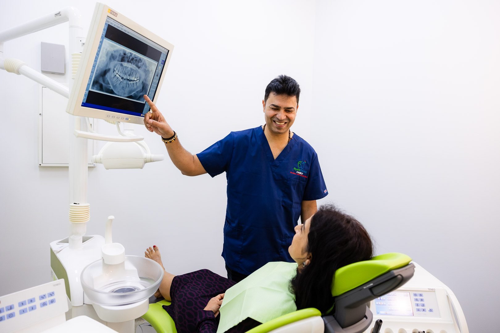 A smiling male dentist or dental technician in navy scrubs pointing at an X-ray of teeth on a monitor, while a woman patient lies in a dental chair with a green headrest in a clean, white dental clinic.
