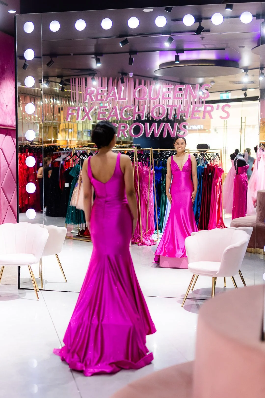 A woman in a pink evening gown looking at her reflection in a mirror inside a dress shop. The shop has a sign that reads 'Real Queens Fix Each Other's Crowns' and displays racks of colorful dresses.