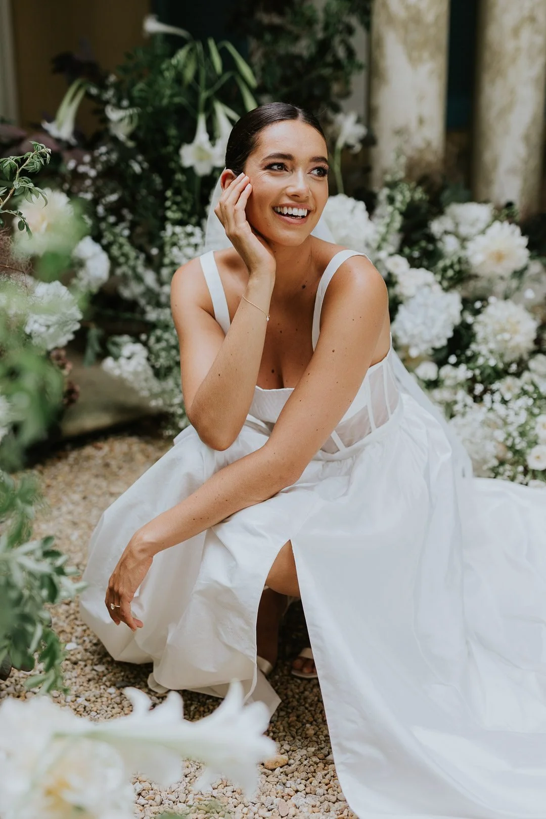 A woman in a white dress sitting outdoors, surrounded by white flowers and greenery, smiling and touching her face.