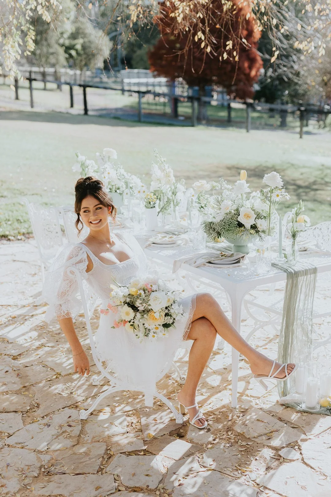 A woman in a white lace dress sitting at an outdoor table decorated with white flowers, holding a bouquet, on a sunny day.