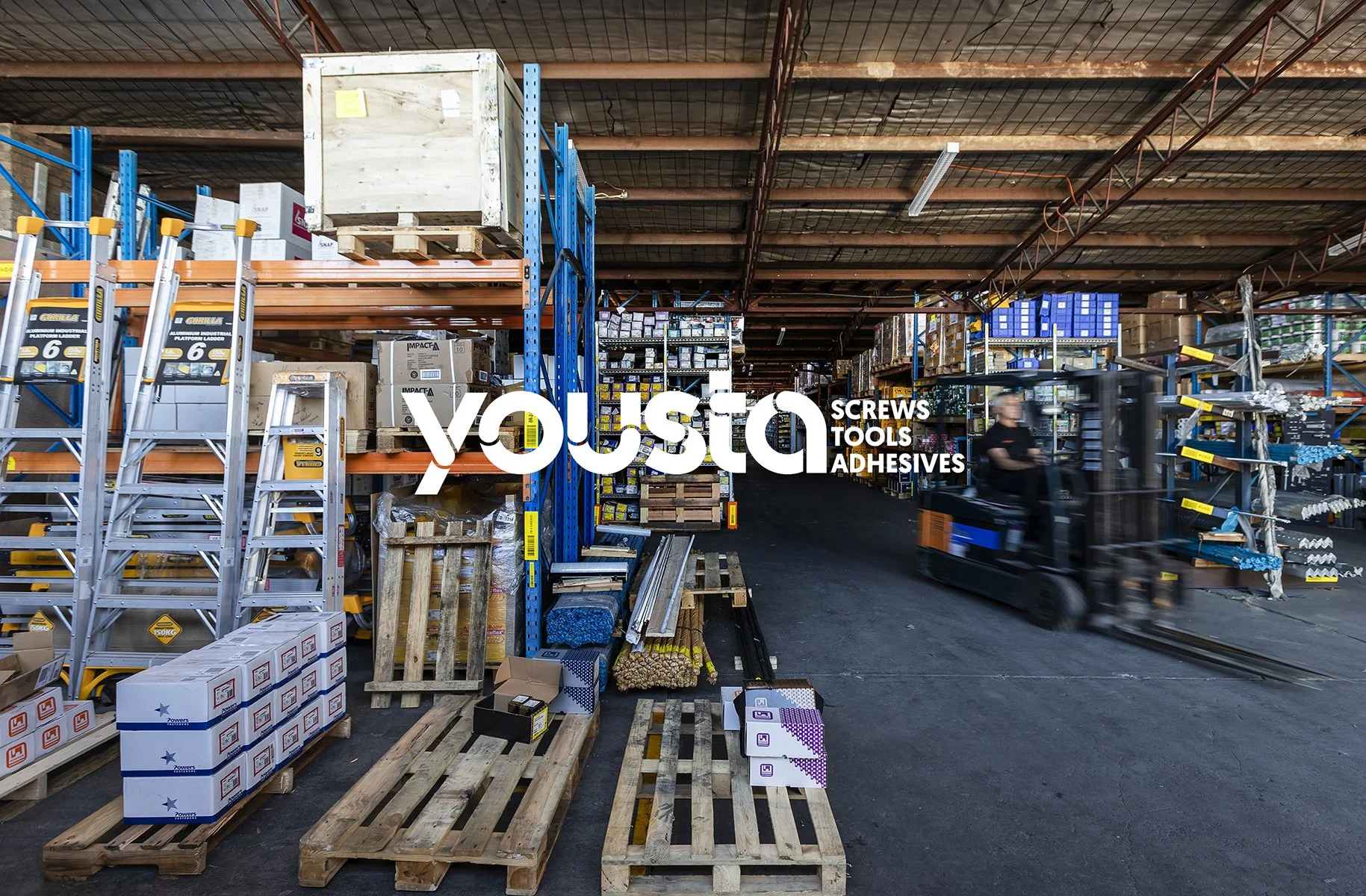The interior of a factory full of construction supplies, with a blurred forklift truck being driven through the shot. In the centre of the image is a company logo which says Yousta, Screws Tools Adhesives. It is a company rebrand photo.