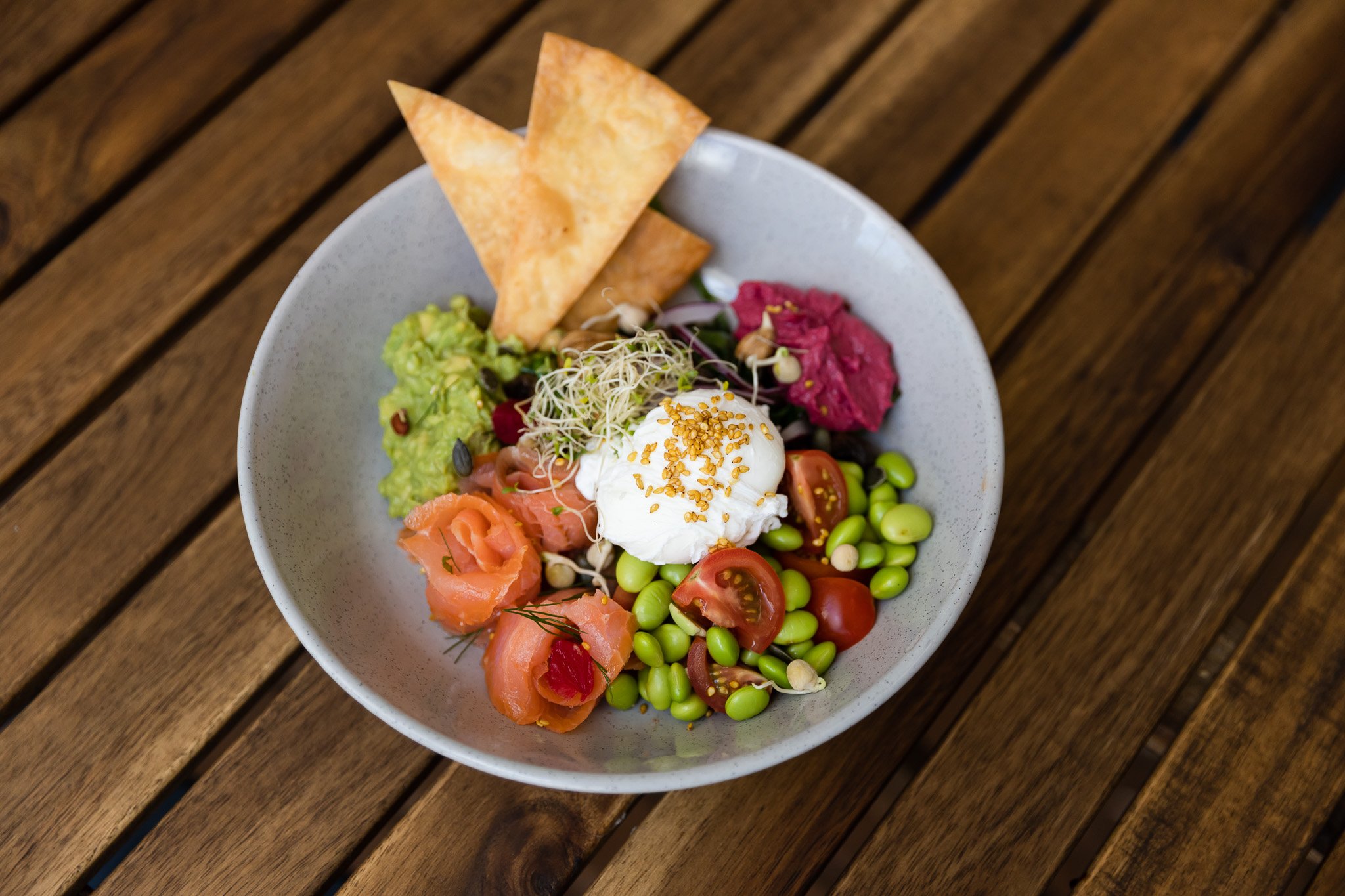 Salad with smoked salmon, cherry tomatoes, edamame, purple beet hummus, guacamole, microgreens, pita chips, and a poached egg, served in a white bowl on a wooden table.