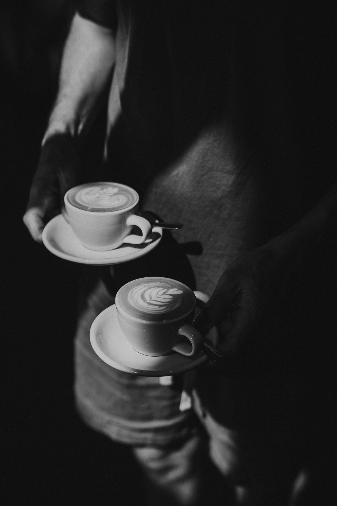 Two people holding cups of coffee with latte art, seen from above, in a black-and-white photo.
