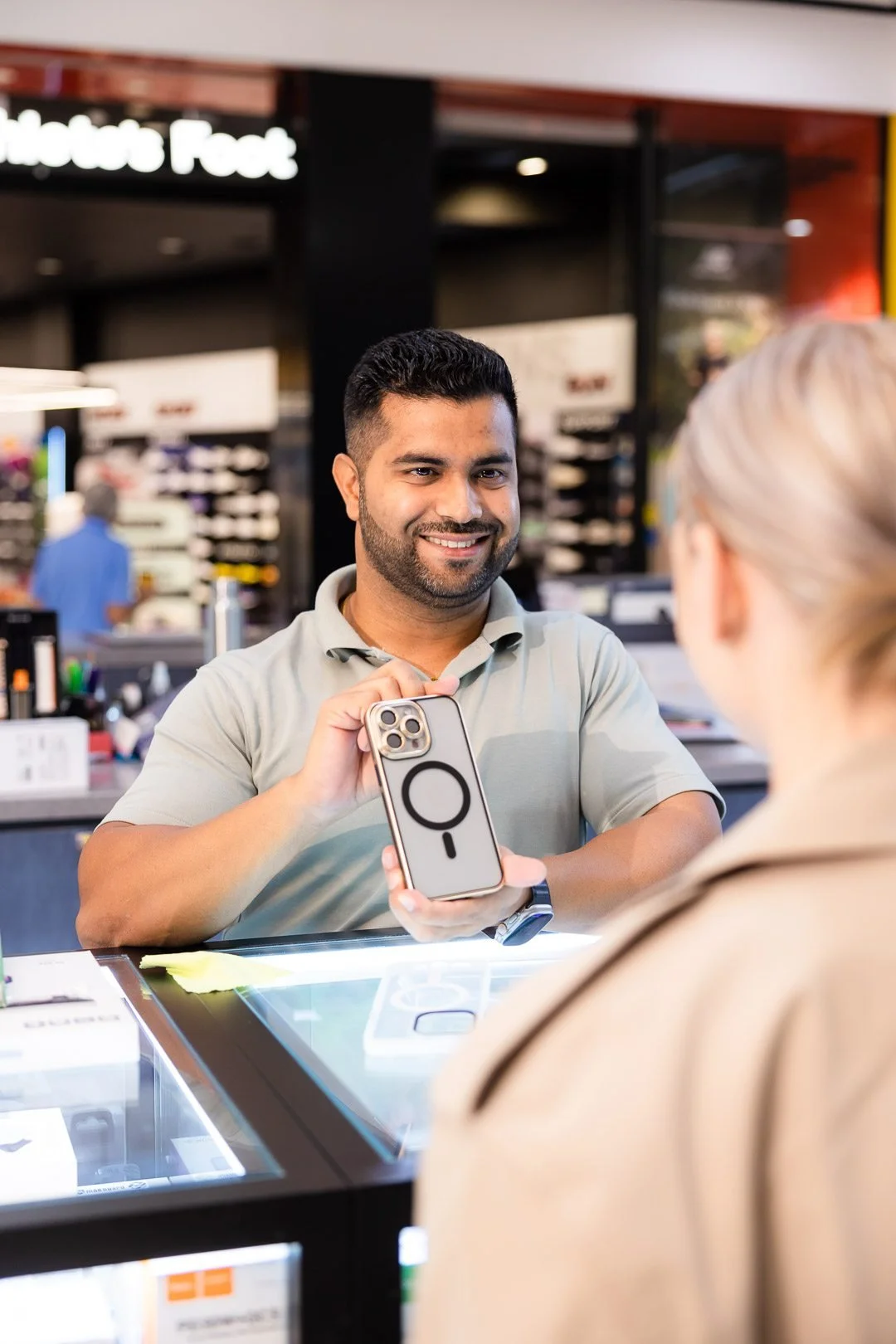 A man with dark hair and a beard smiling at a woman while holding a smartphone with a pop socket at a retail counter.