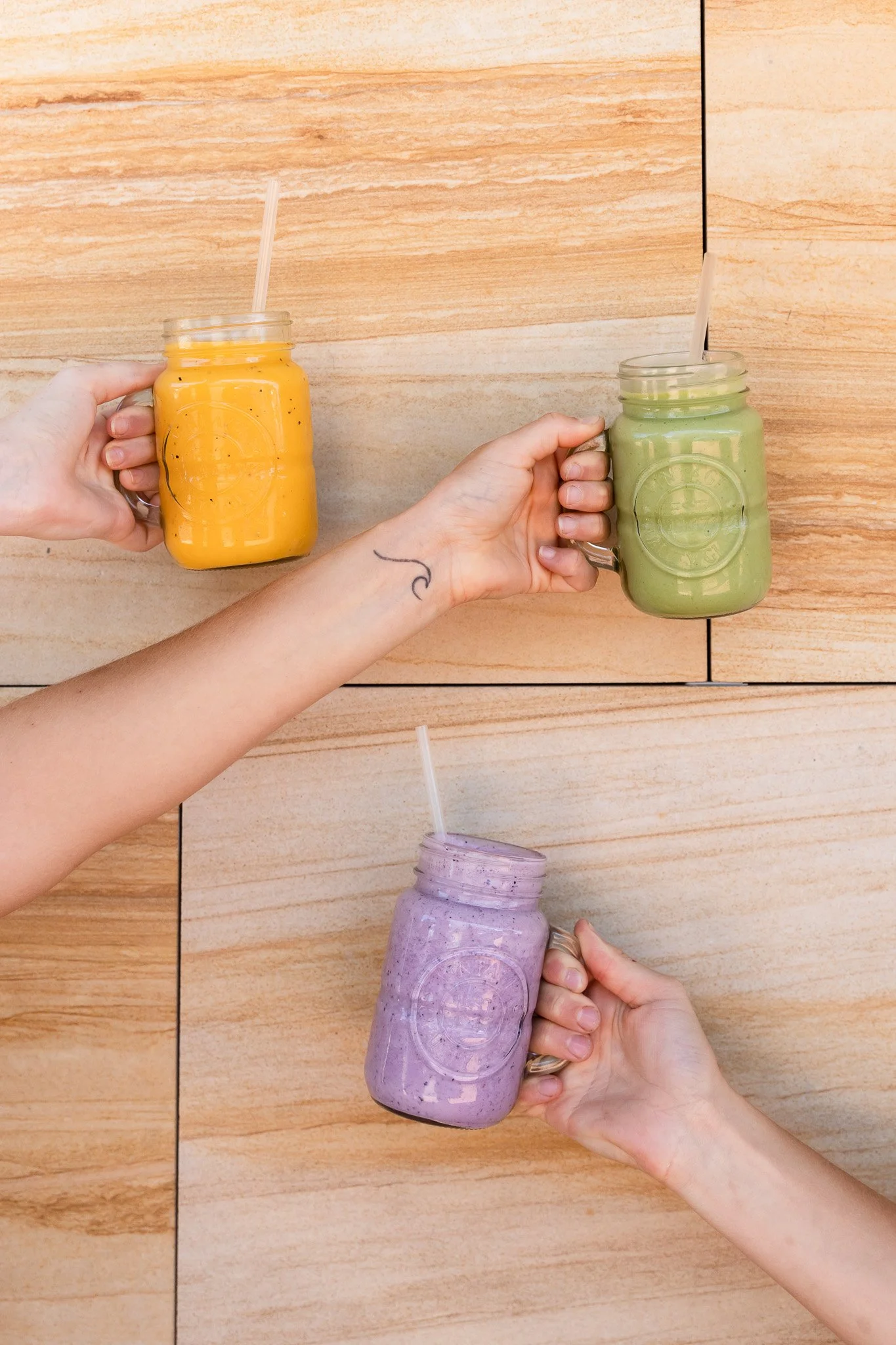Three hands holding mason jars filled with colorful smoothies on a wooden tiled surface.