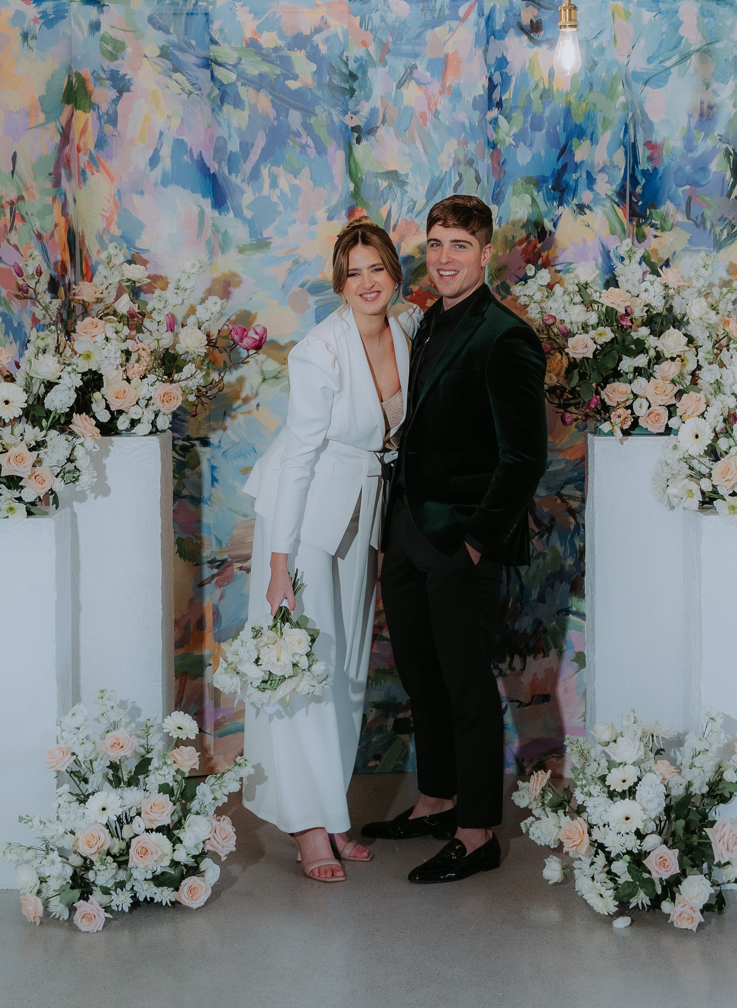 A couple at their wedding, standing together in front of a colorful floral backdrop surrounded by white and blush flower arrangements on pedestals. The bride is in a white suit with a bouquet and the groom is in a black velvet blazer and black trouse