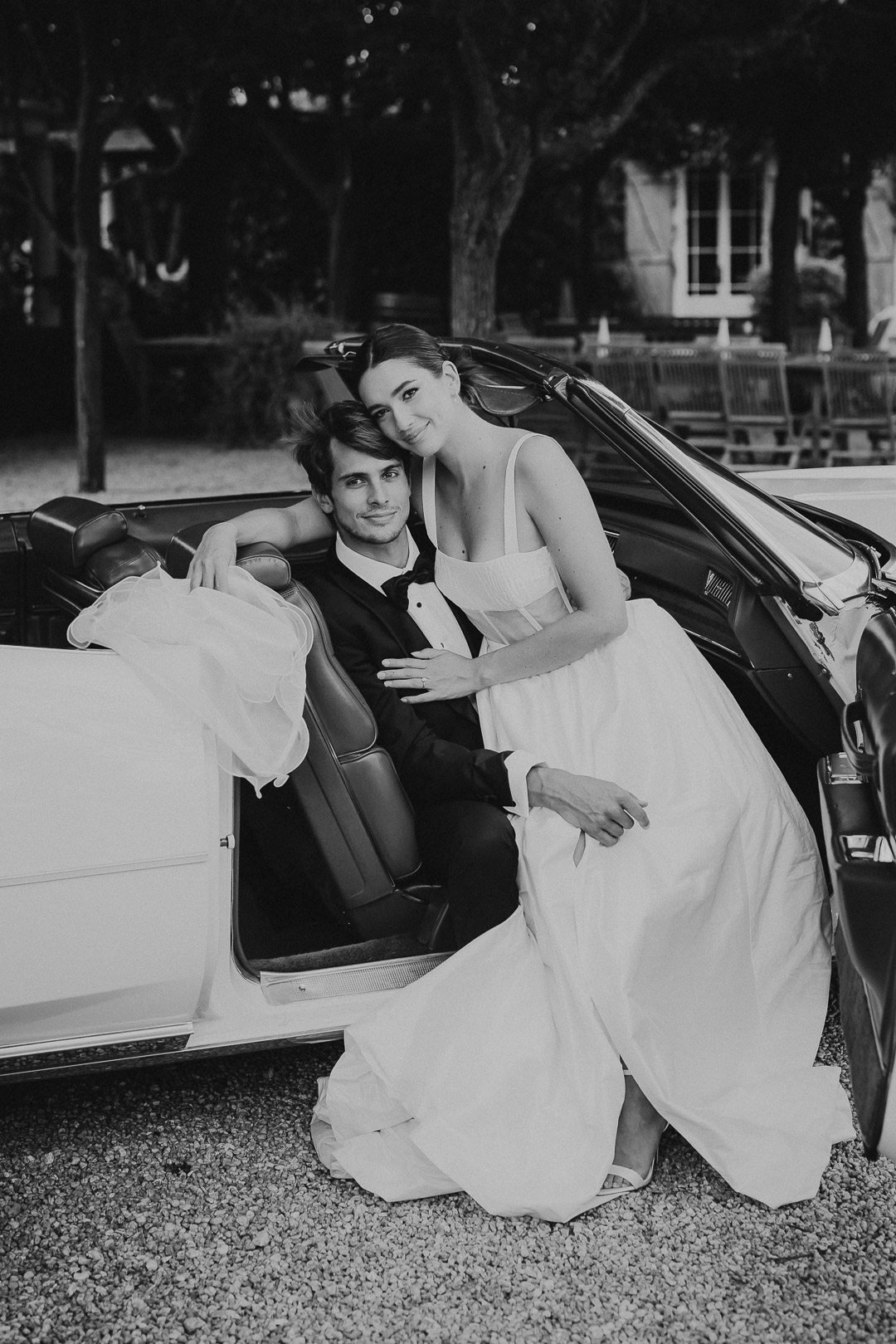 Black-and-white photo of a bride and groom sitting in a convertible car, with the bride leaning on the groom's shoulder outdoors, surrounded by trees and benches.