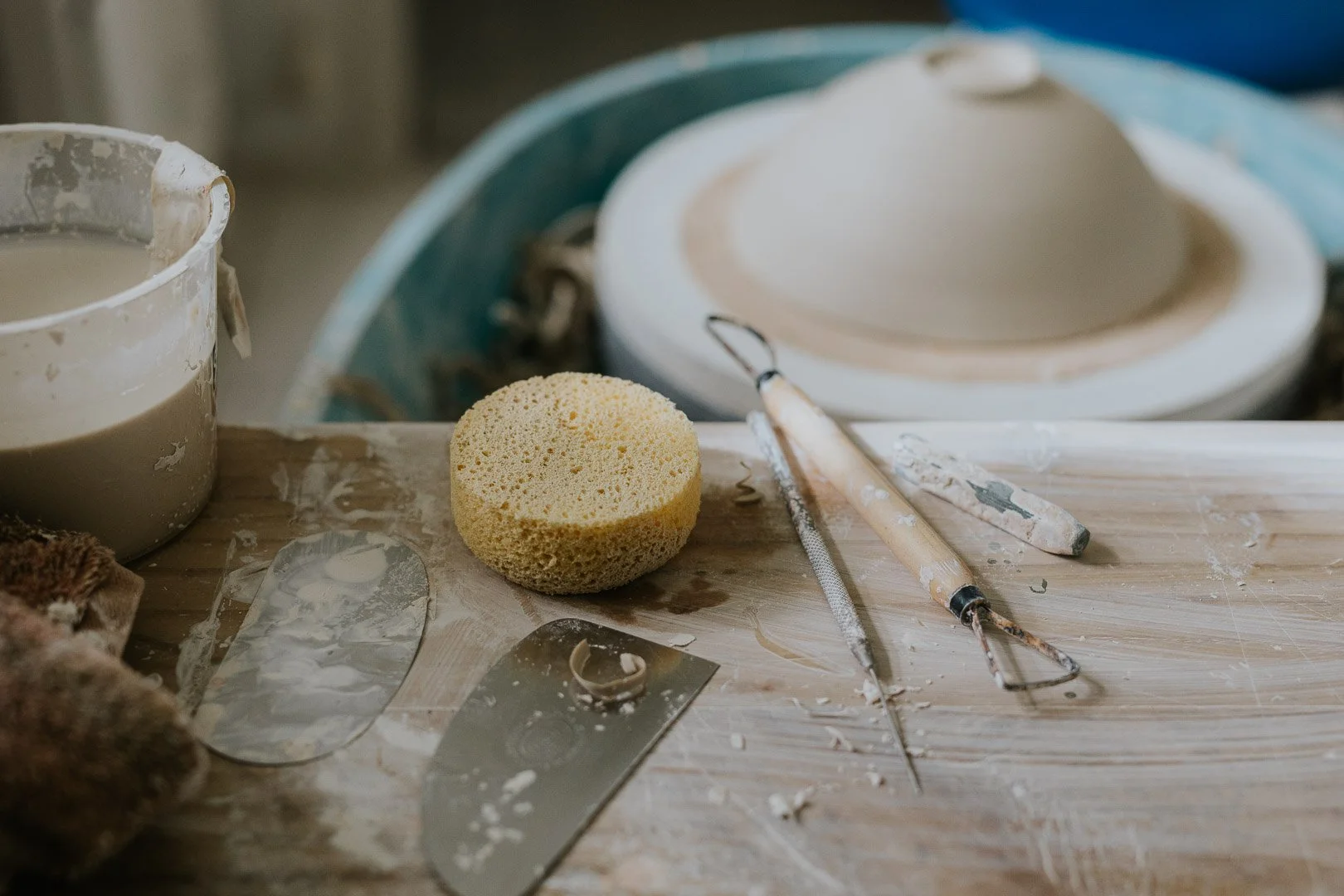 Pottery tools and supplies on a worktable, including a sponge, palette knife, carving tools, clay, and a spinning pottery wheel in the background.