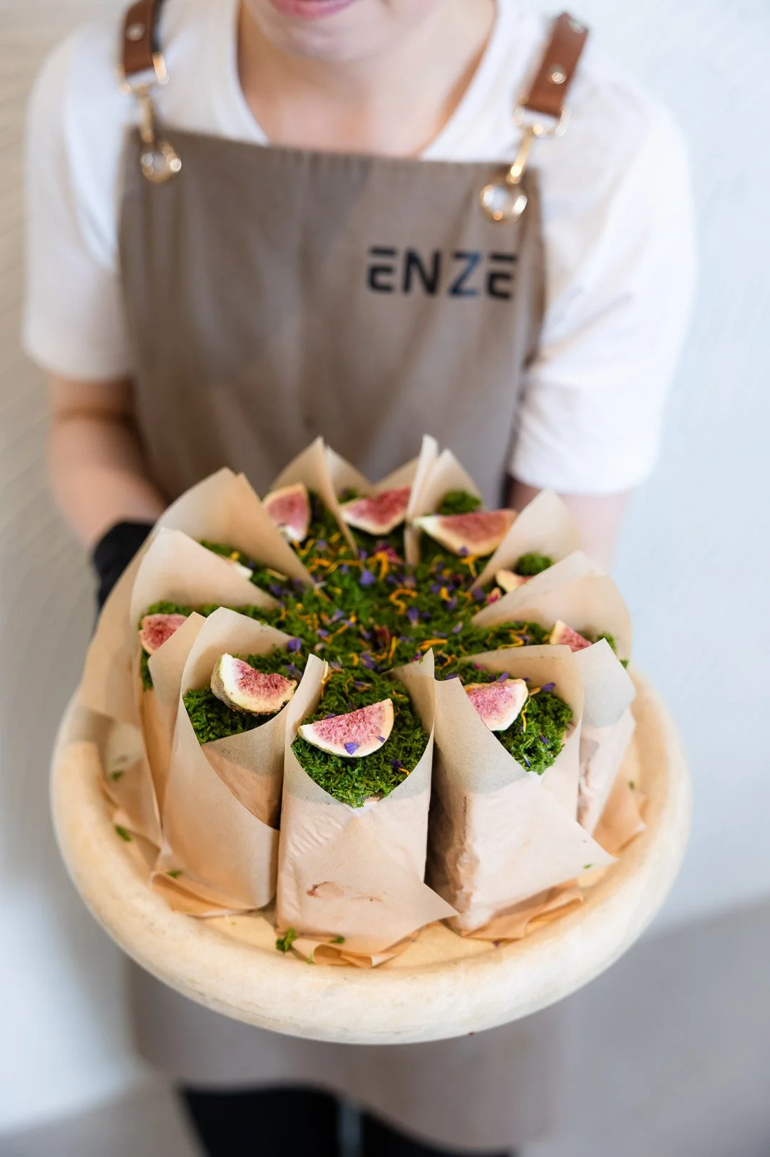 A person wearing an apron holding a large round tray with rolled sandwiches topped with fig slices and edible flowers.