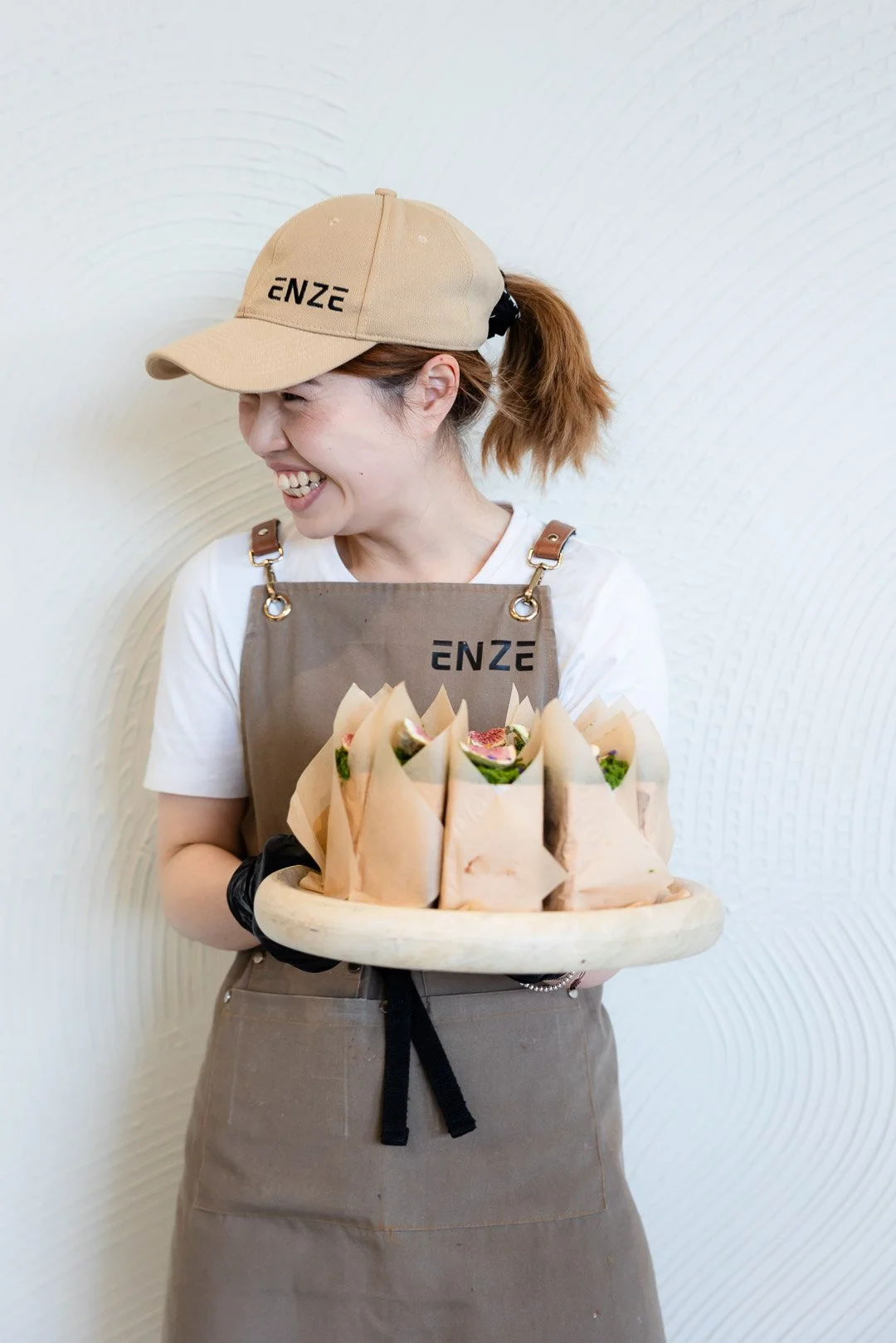 A woman wearing a beige cap, tan apron with the brand 'ENZE,' smiling and holding a tray of wrapped sandwiches or wraps in front of a white textured wall.