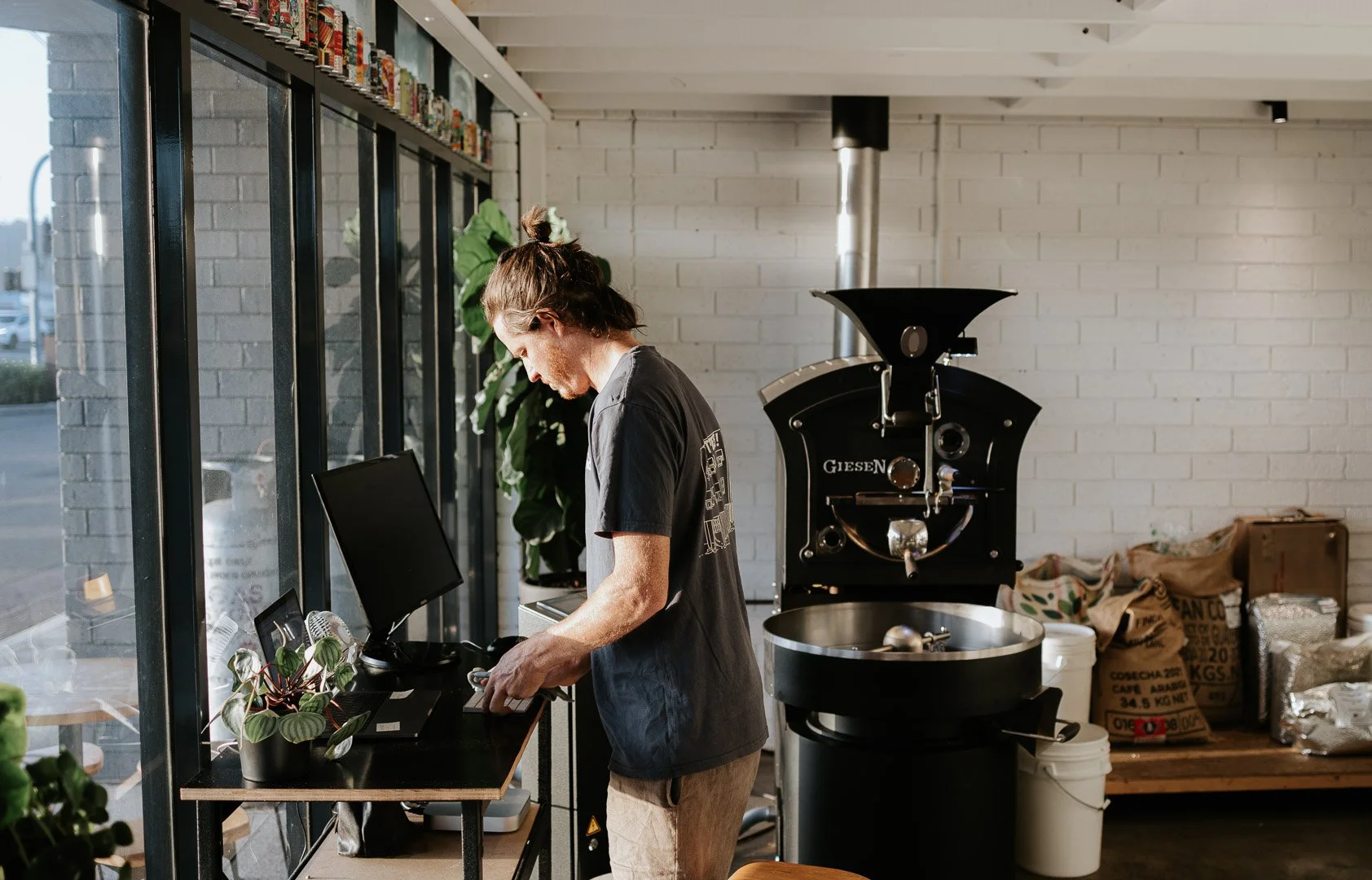 Natural documentary branding photography for small businesses on the Central Coast. A man is working at a small desk with a computer and scale inside a cafe, with a large coffee roaster machine nearby.