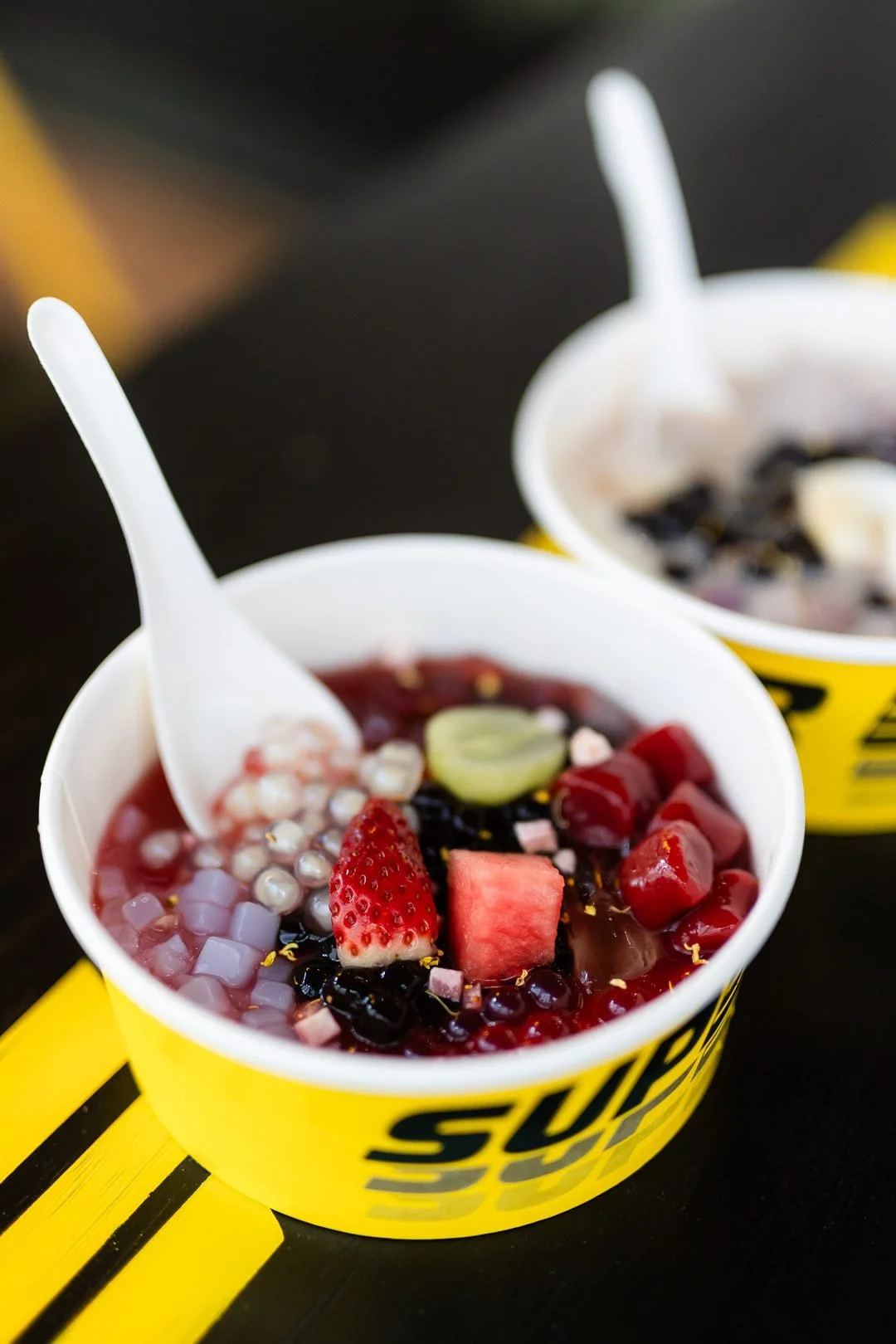 Two cups of dessert with colorful toppings, including fruits and jelly, placed on a black and yellow surface.