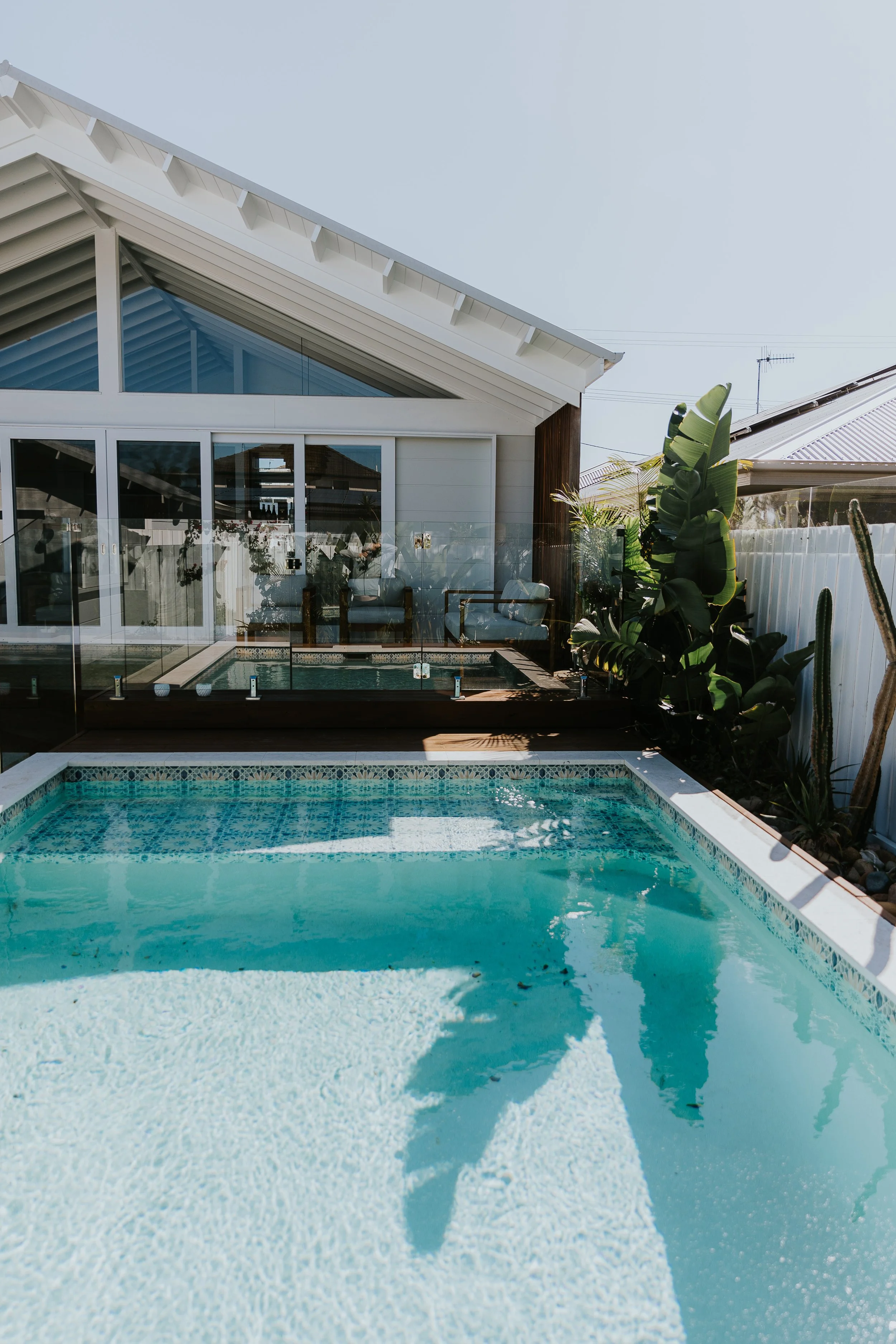 A backyard scene with a swimming pool in the foreground, a fenced patio area with glass barriers, outdoor seating, and a modern house with large glass doors and a white sloped roof in the background. Green plants and cacti are along the fence.