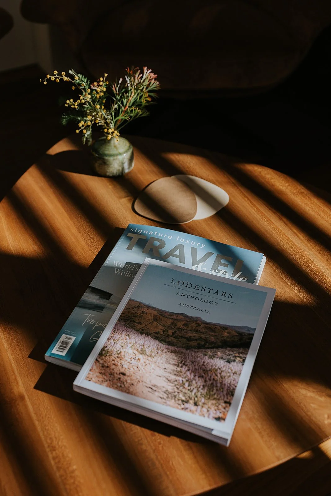 Editorial Brand Imagery Sydney. A wooden table holding a small vase with yellow flowers, two magazines, and a rock. Sunlight creates shadows on the table.