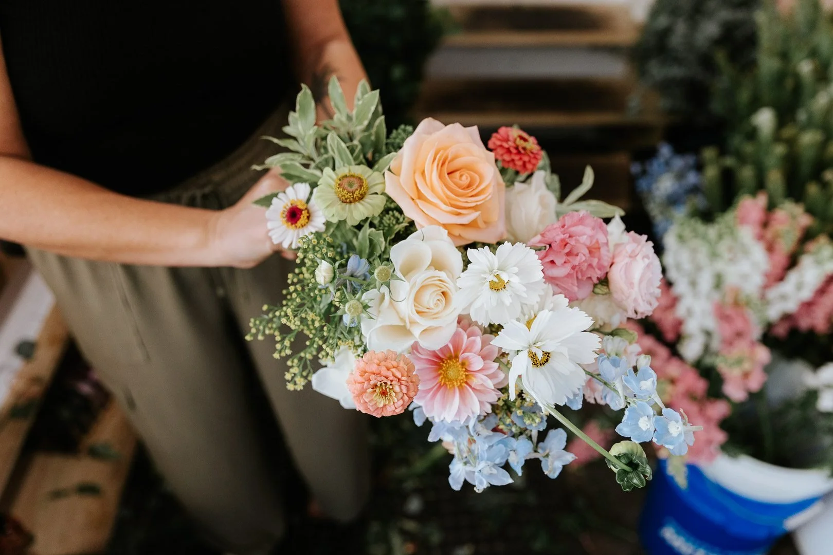 Person holding a bouquet of mixed flowers including roses, daisies, hydrangeas, and other blossoms in pastel colors.