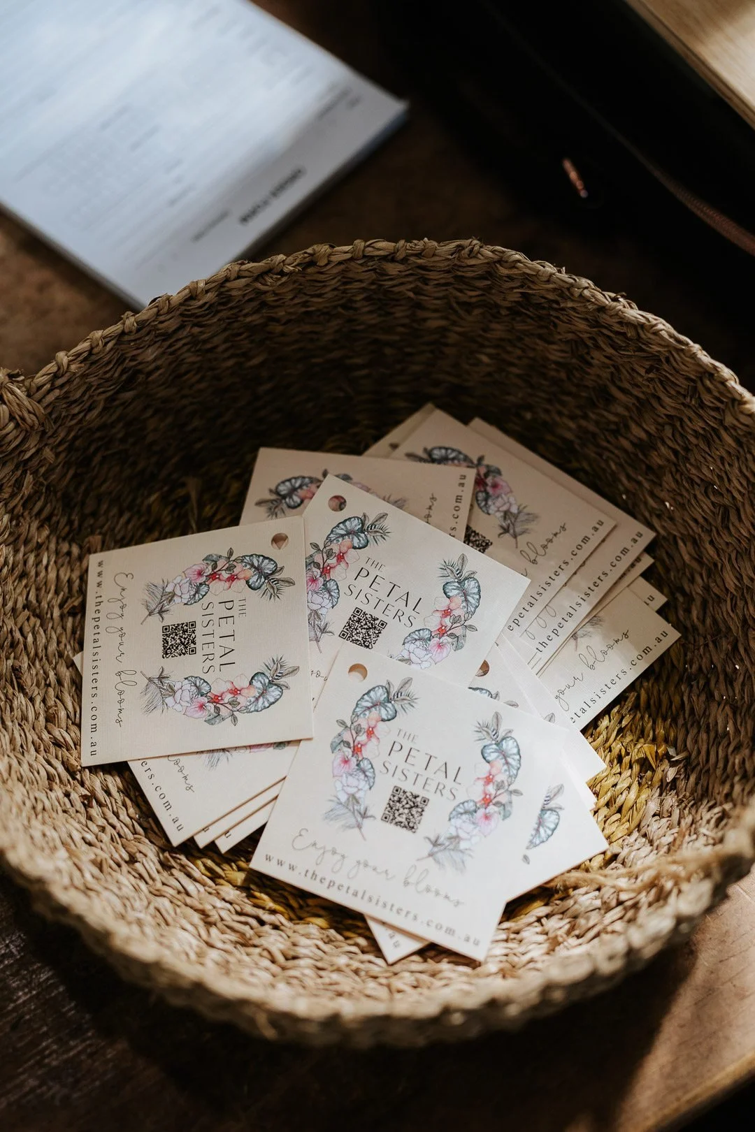 A woven basket filled with business cards for The Petal Sisters, featuring floral illustrations and a QR code, on a wooden surface.