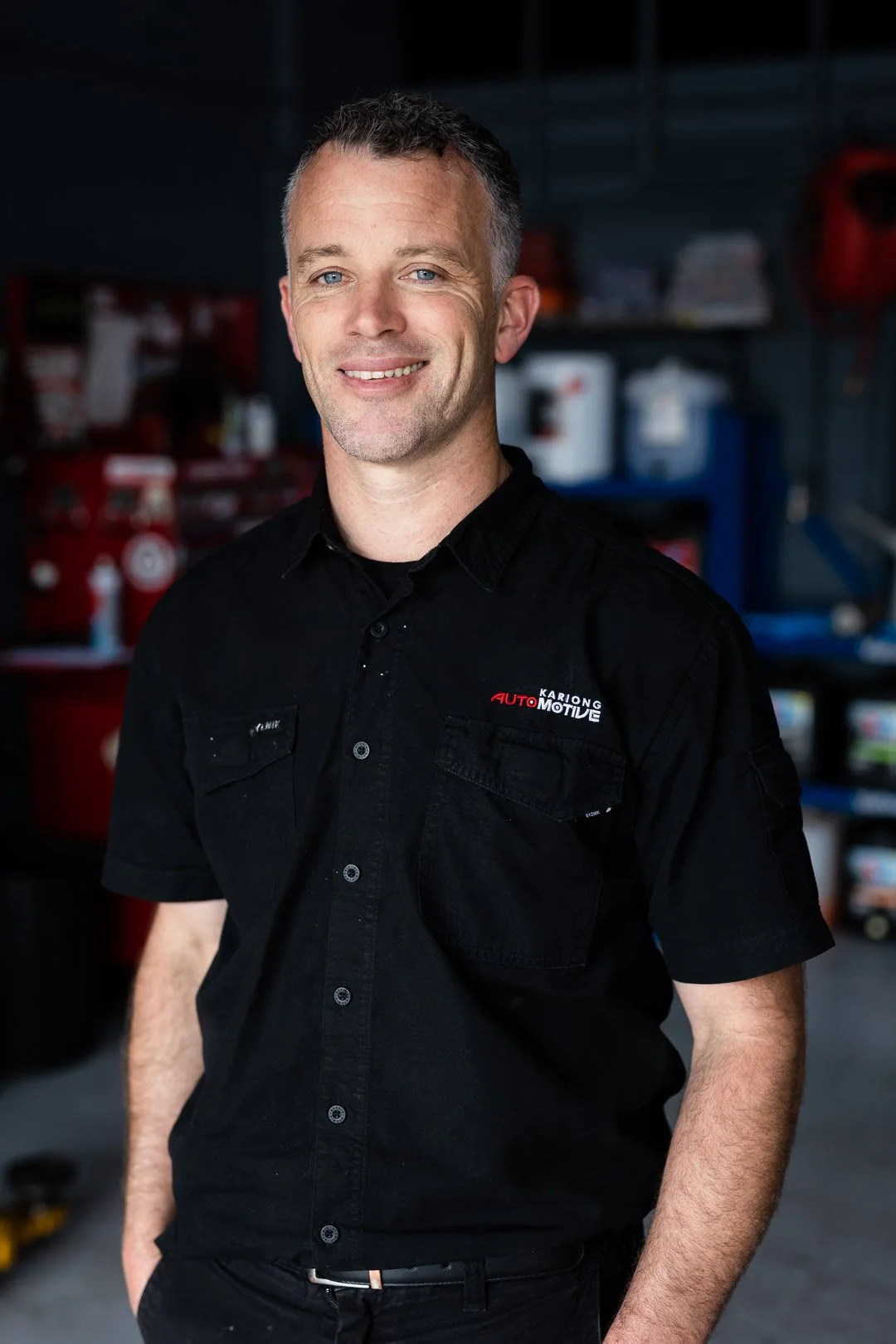 Man smiling in a workshop, wearing a black uniform with 'Kariong Automotive' embroidered on it.