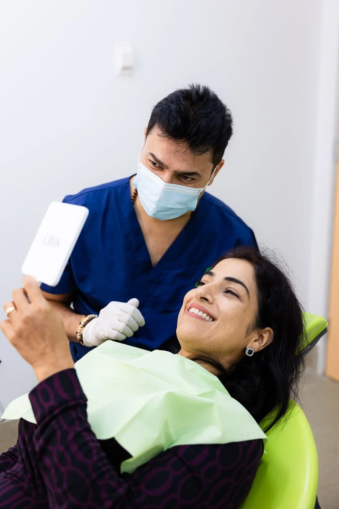 A woman lying in a dental chair smiling at a dentist wearing a surgical mask and gloves. The woman appears relaxed and happy.