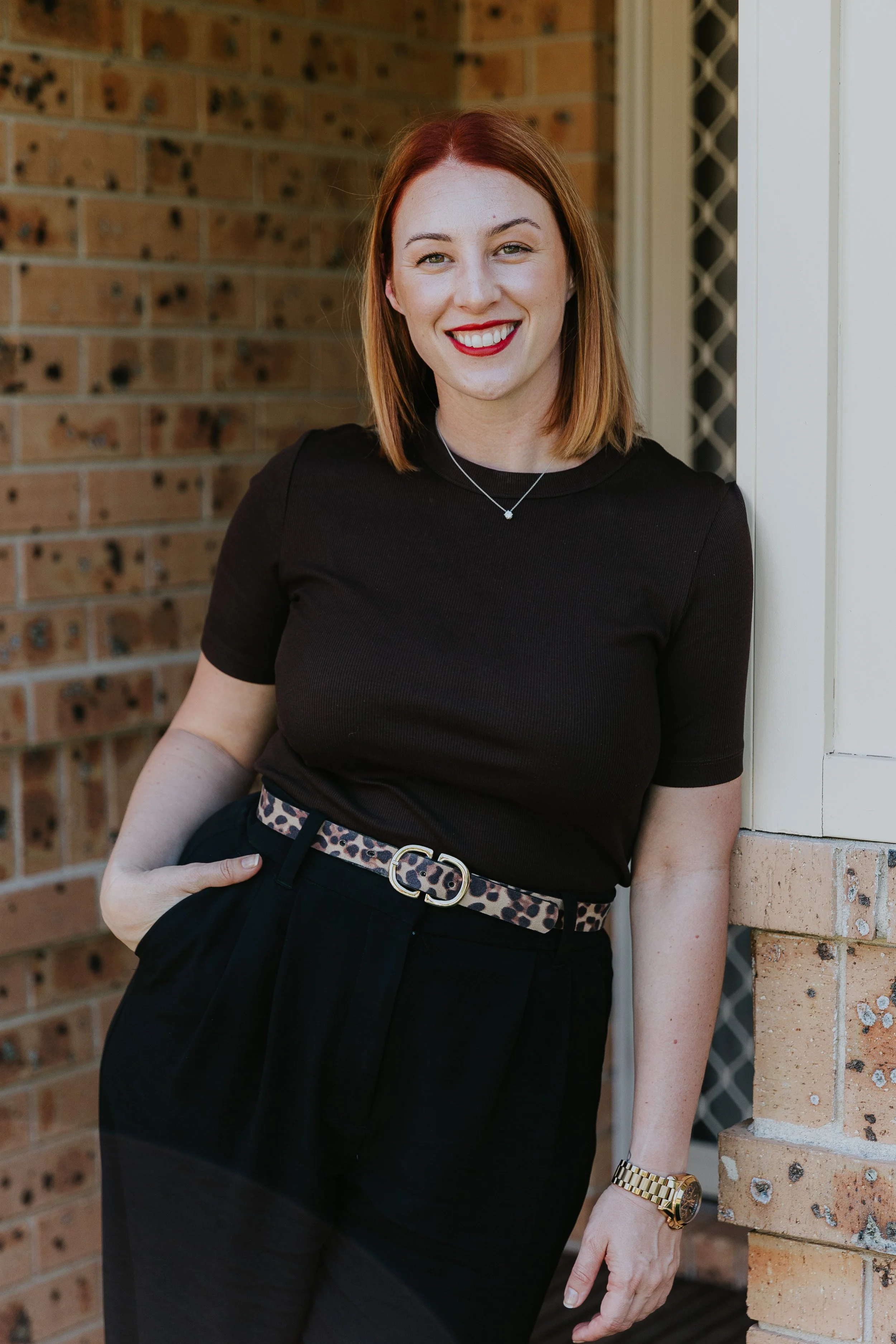 A woman with shoulder-length red hair and red lipstick smiling, wearing a black top, black pants, a leopard print belt, a gold watch, and a necklace, standing outdoors against a brick wall.