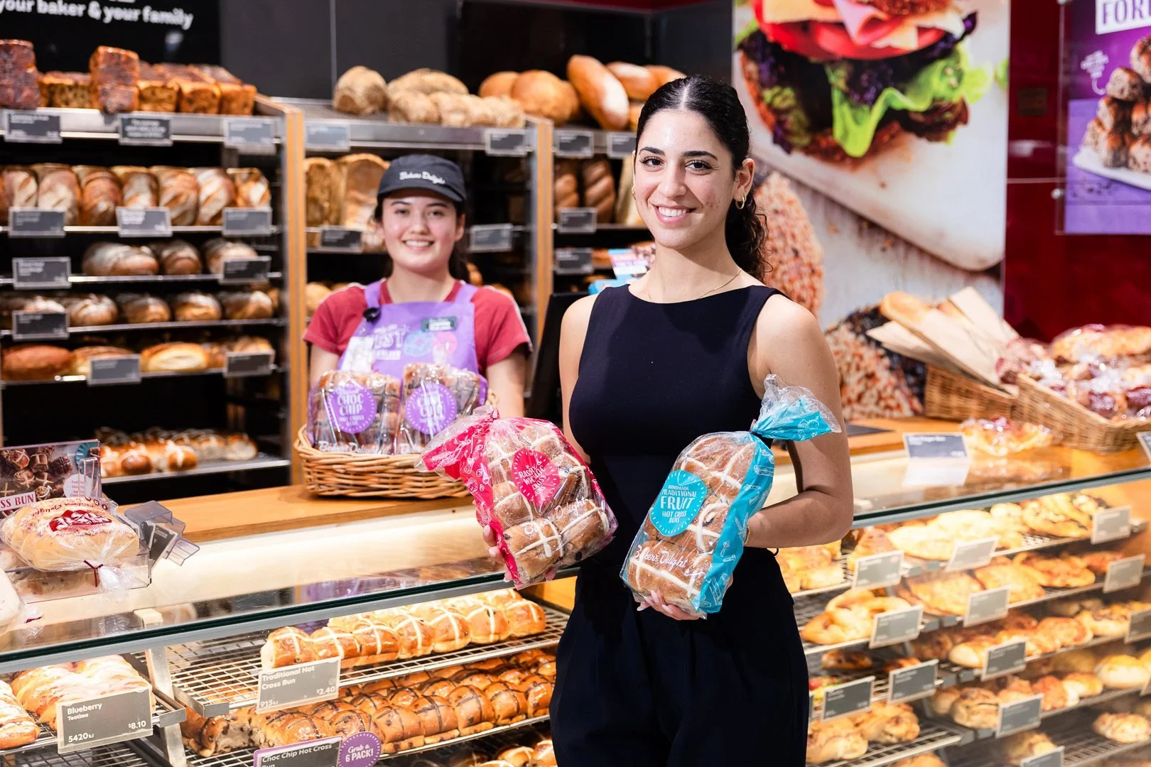 Two women shopping for bread at a bakery section in a grocery store. The woman in front is holding two loaves of bread, smiling, and wearing a black sleeveless dress. The woman behind her is also smiling, wearing a red shirt, purple apron, and a blac