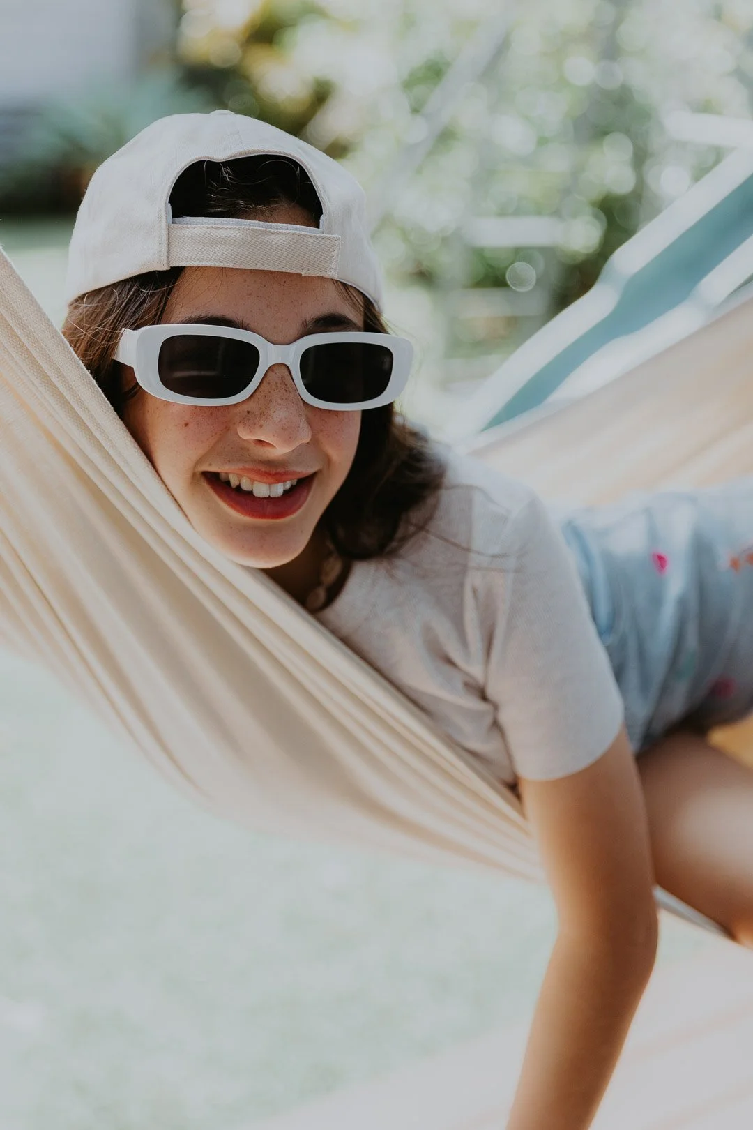 A young woman lying in a hammock, wearing a white baseball cap and white sunglasses, smiling outdoors with greenery in the background.