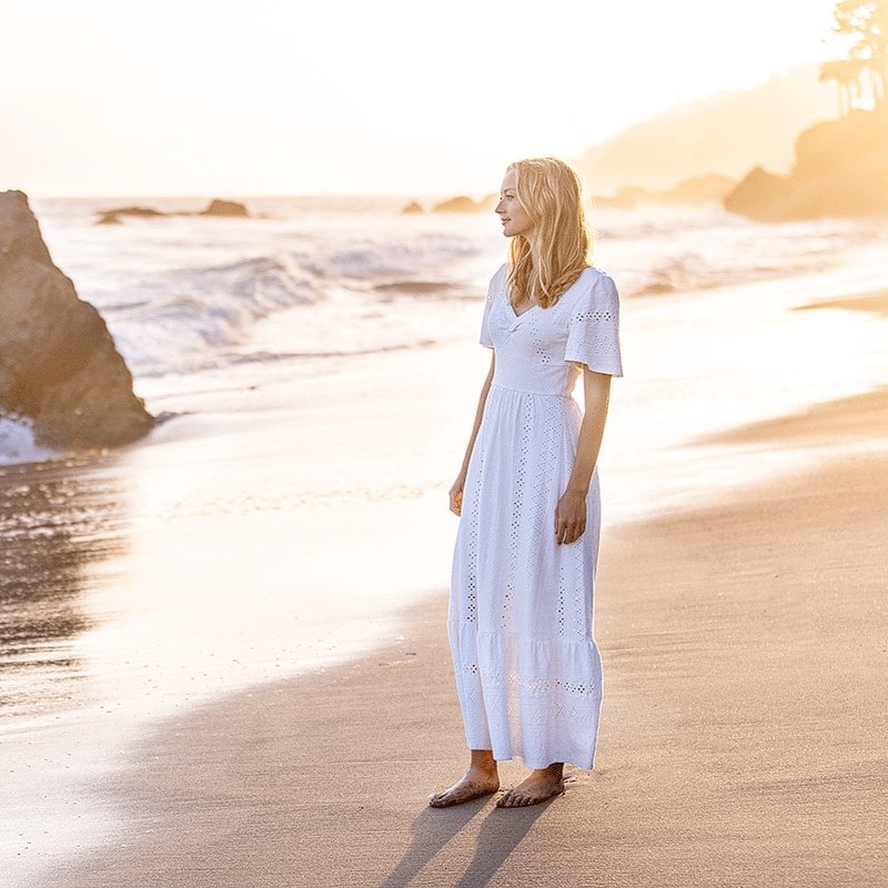 A woman in a white dress standing barefoot on the beach during sunset during a senior portrait photo shoot, with ocean waves and rocky formations in the background.