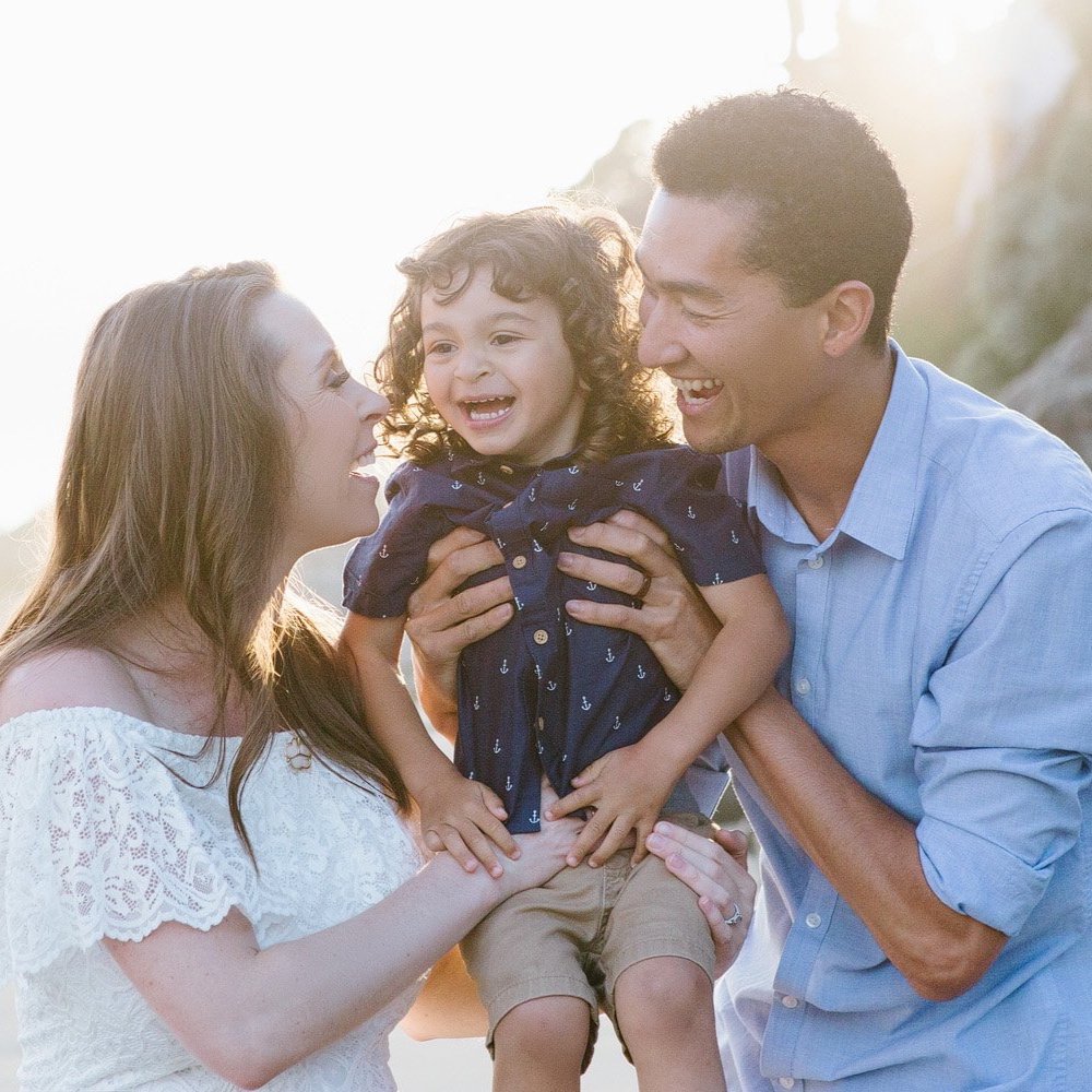 A happy family of three smiling and playing outdoors in the sunlight, holding a young boy between them.