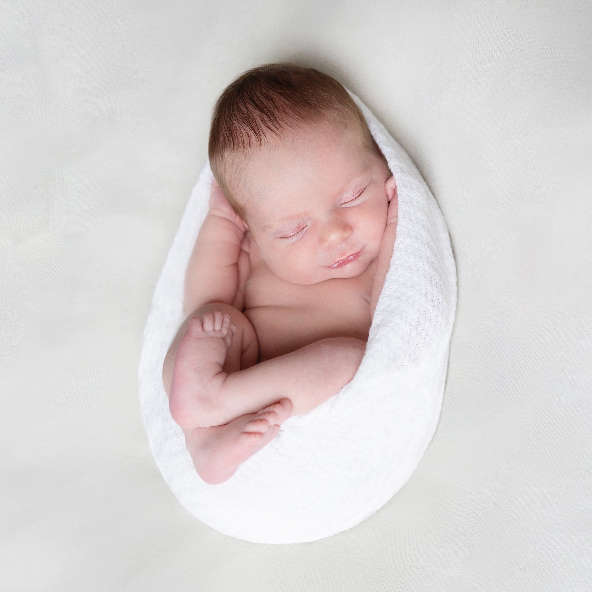 A newborn baby wrapped in a white blanket for a newborn photo shoot, sleeping peacefully with hands near face, on a light background.