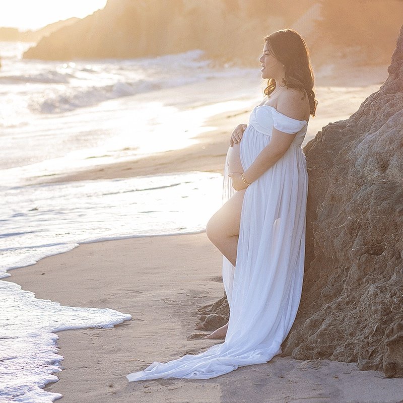 Pregnant woman in a white dress leaning against a rock on the beach at sunset during a maternity photo session..