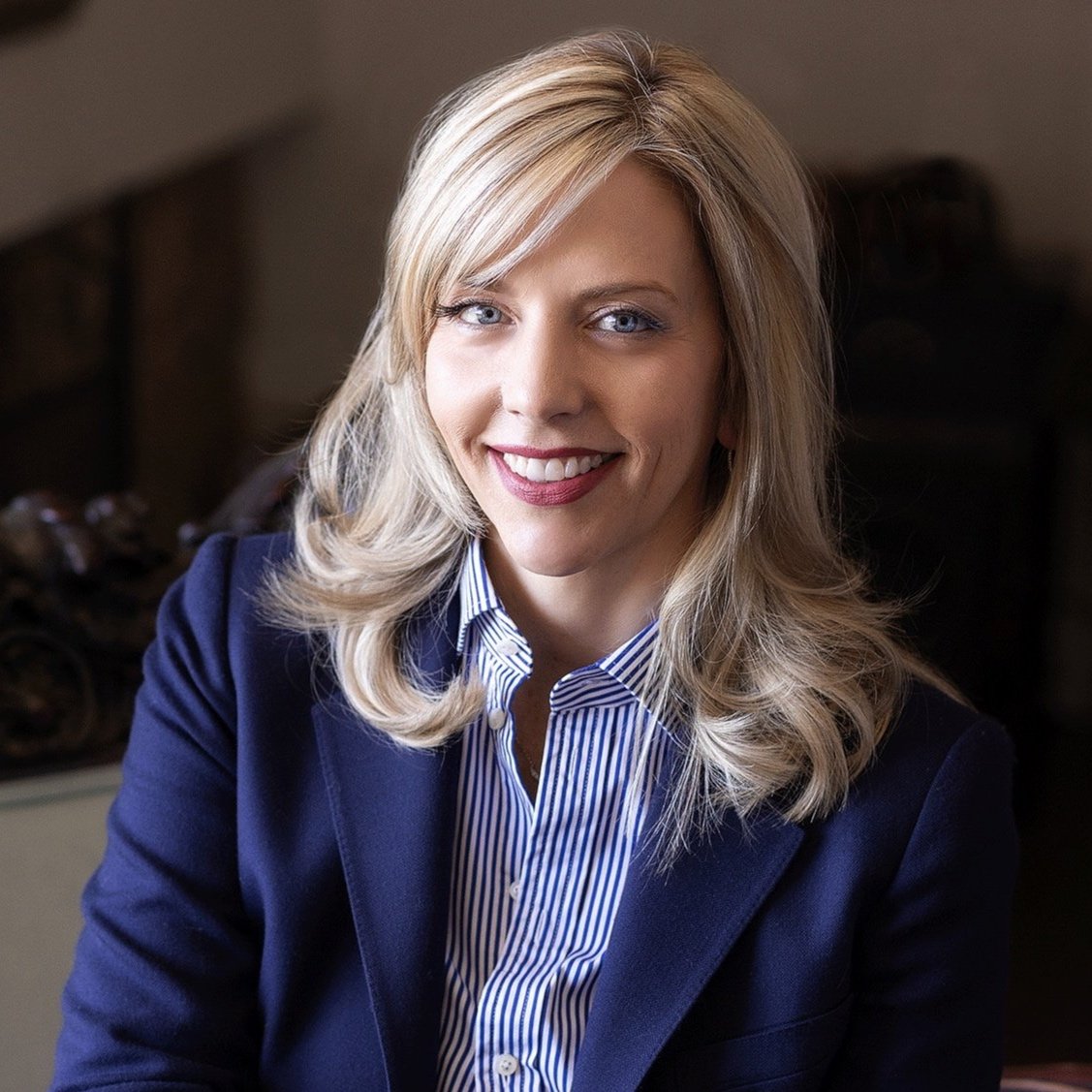 A woman with long blonde hair and blue eyes, wearing a dark blue blazer and a striped shirt, smiling at the camera.