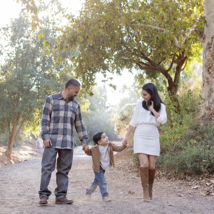 A family of three walking hand in hand on a dirt trail in a park, surrounded by trees with green leaves during a family photo shoot.