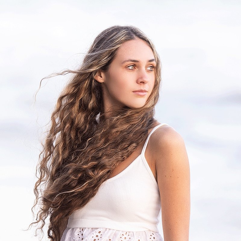 A young woman with long, wavy brown hair wearing a white sleeveless top standing outdoors against a cloudy sky during a senior portrait session.