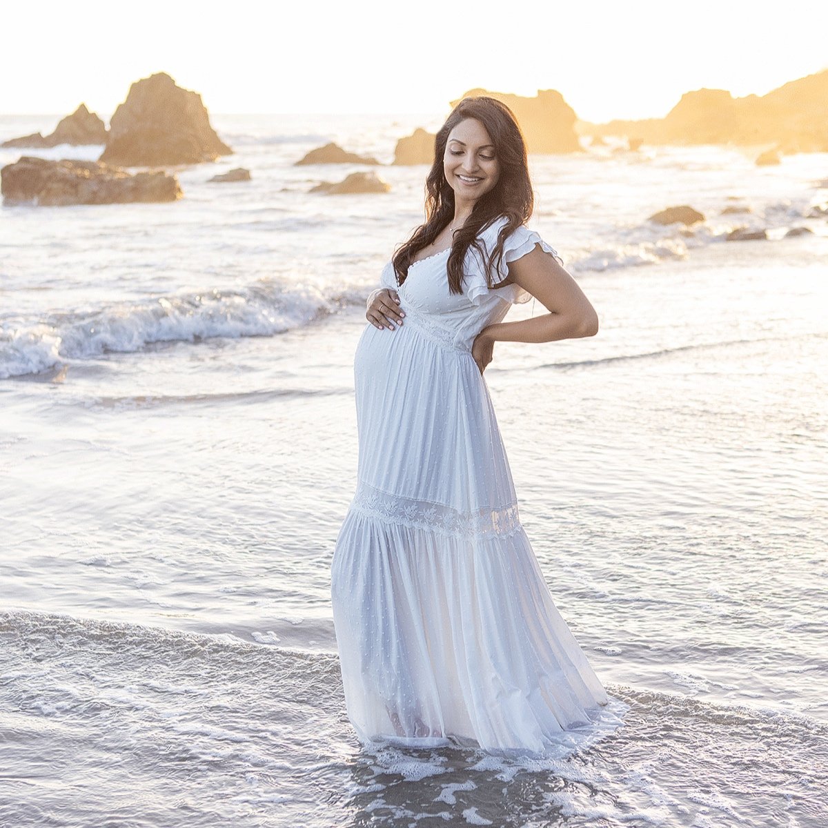 A pregnant woman in a white flowing dress standing in the shallow water of a beach at sunset, smiling and cradling her belly to celebrate her maternity photo shoot