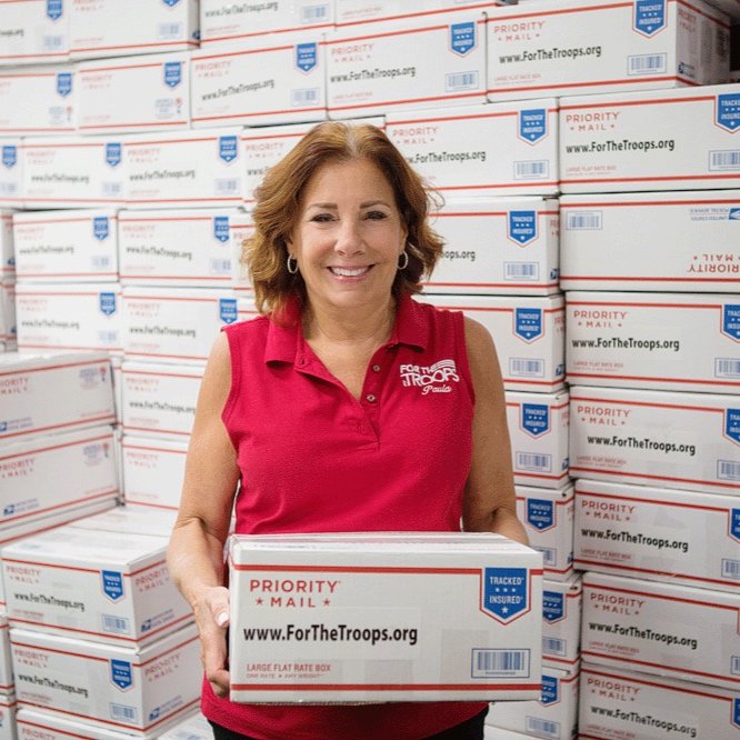 A smiling woman wearing a red sleeveless polo shirt holds a Priority Mail box with the website www.ForTheTroops.org in front of a wall stacked with similar boxes.