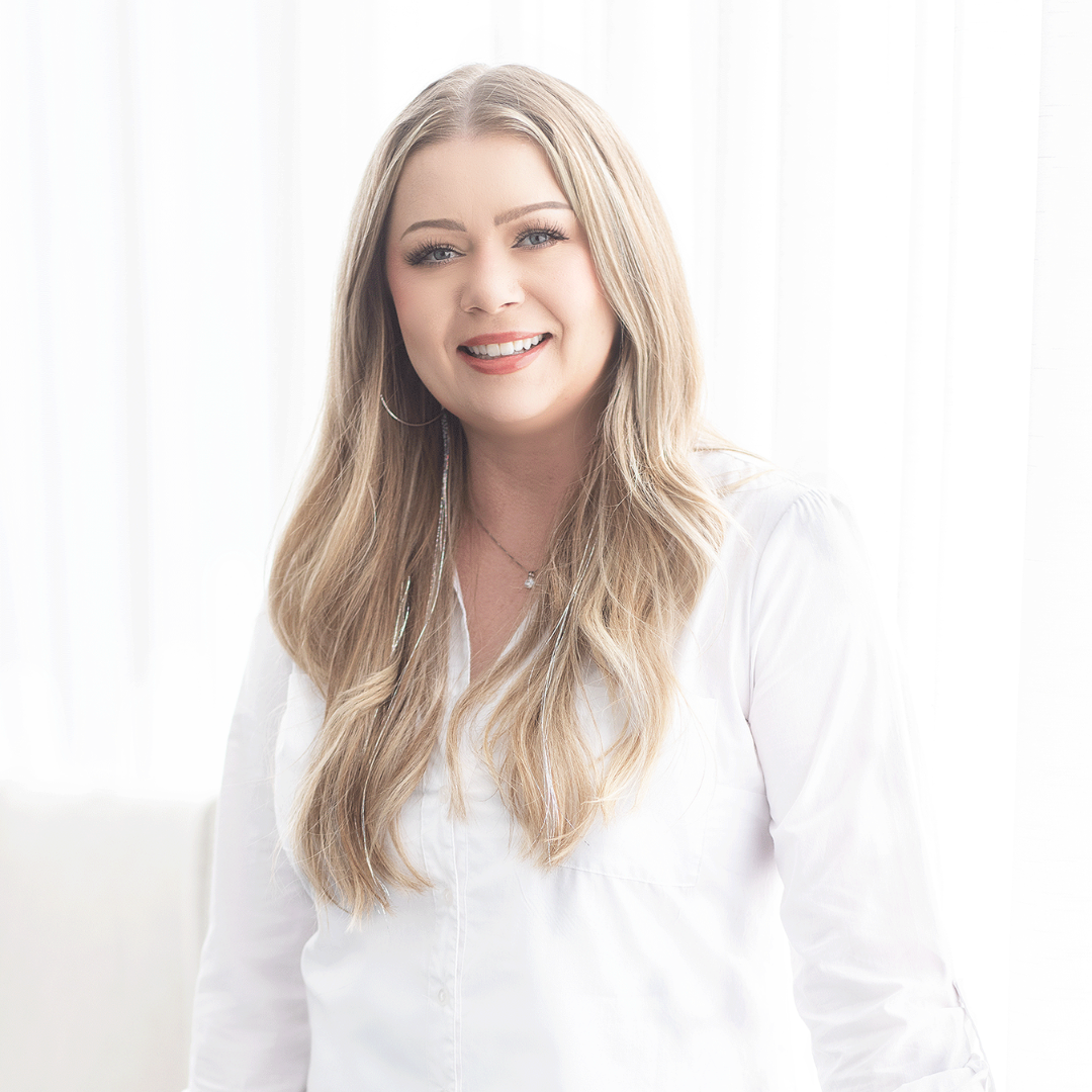 A smiling woman with long blonde hair, wearing a white shirt, standing in front of a bright, white background for a headshot session.