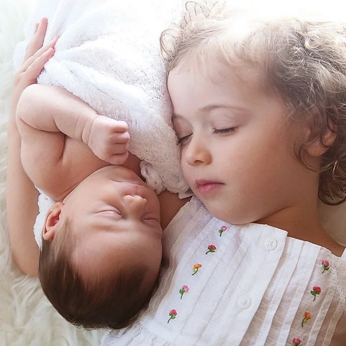 A young girl peacefully sleeping next to a newborn baby, both lying on a soft surface in a newborn photography session.