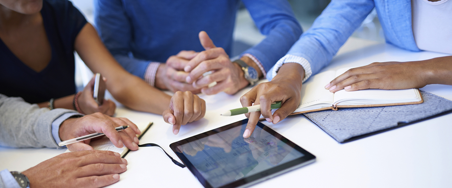Hands of a team of people collaborating using a variety of media and devices, including a notebook, tablet, phone, and map.