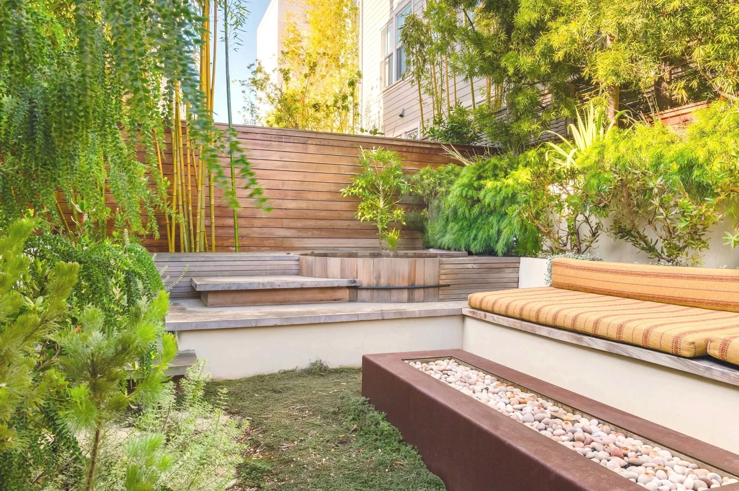 A well-maintained backyard with wooden decking, lush green plants and bushes, a built-in bench with striped cushion, a rectangular planter with white pebbles, and a tall wooden privacy fence, illuminated by sunlight.