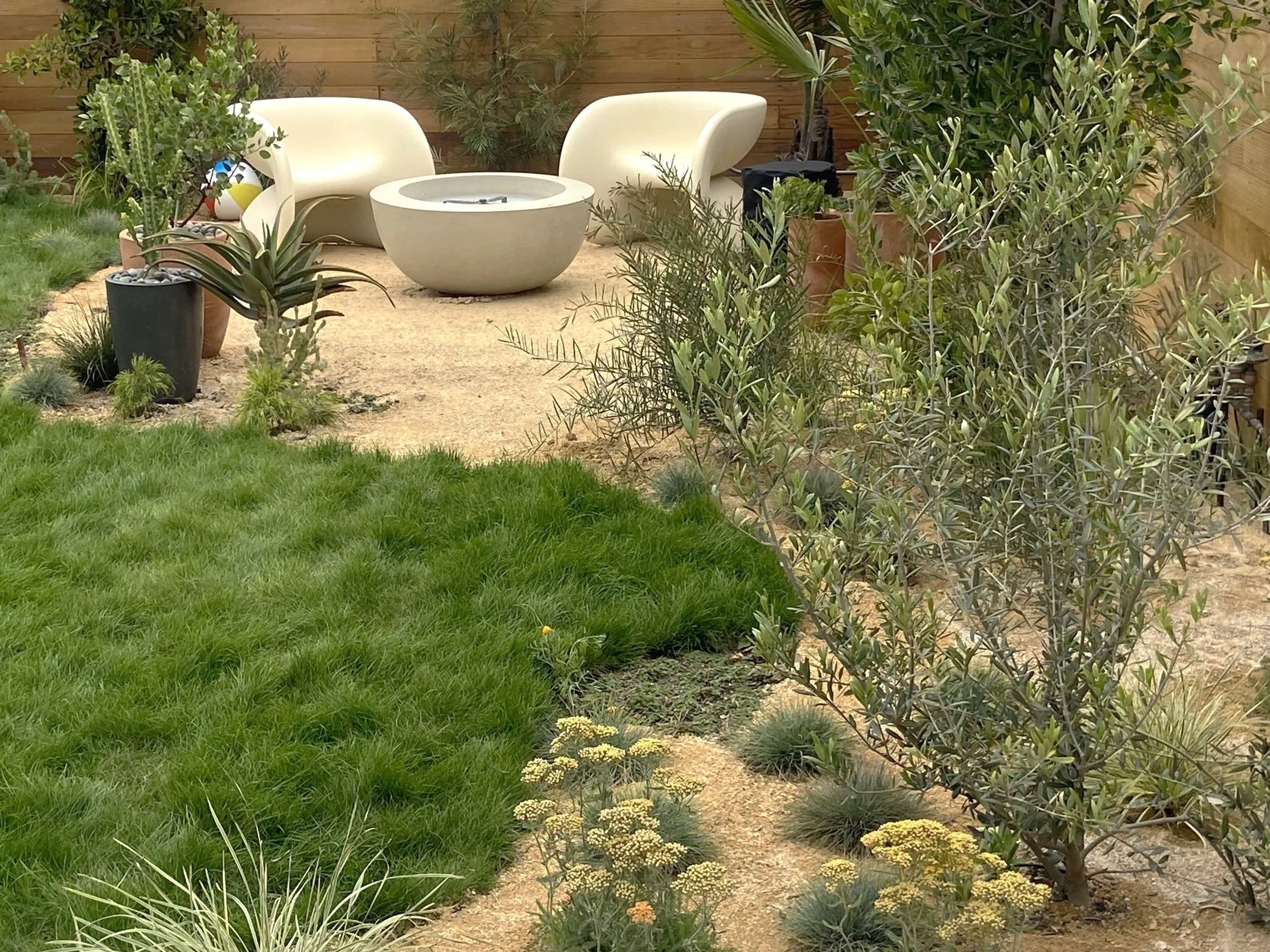 A backyard garden with a grassy patch, potted plants, sandy paths, outdoor seating with chairs, a round table, and various desert plants against a wooden fence.