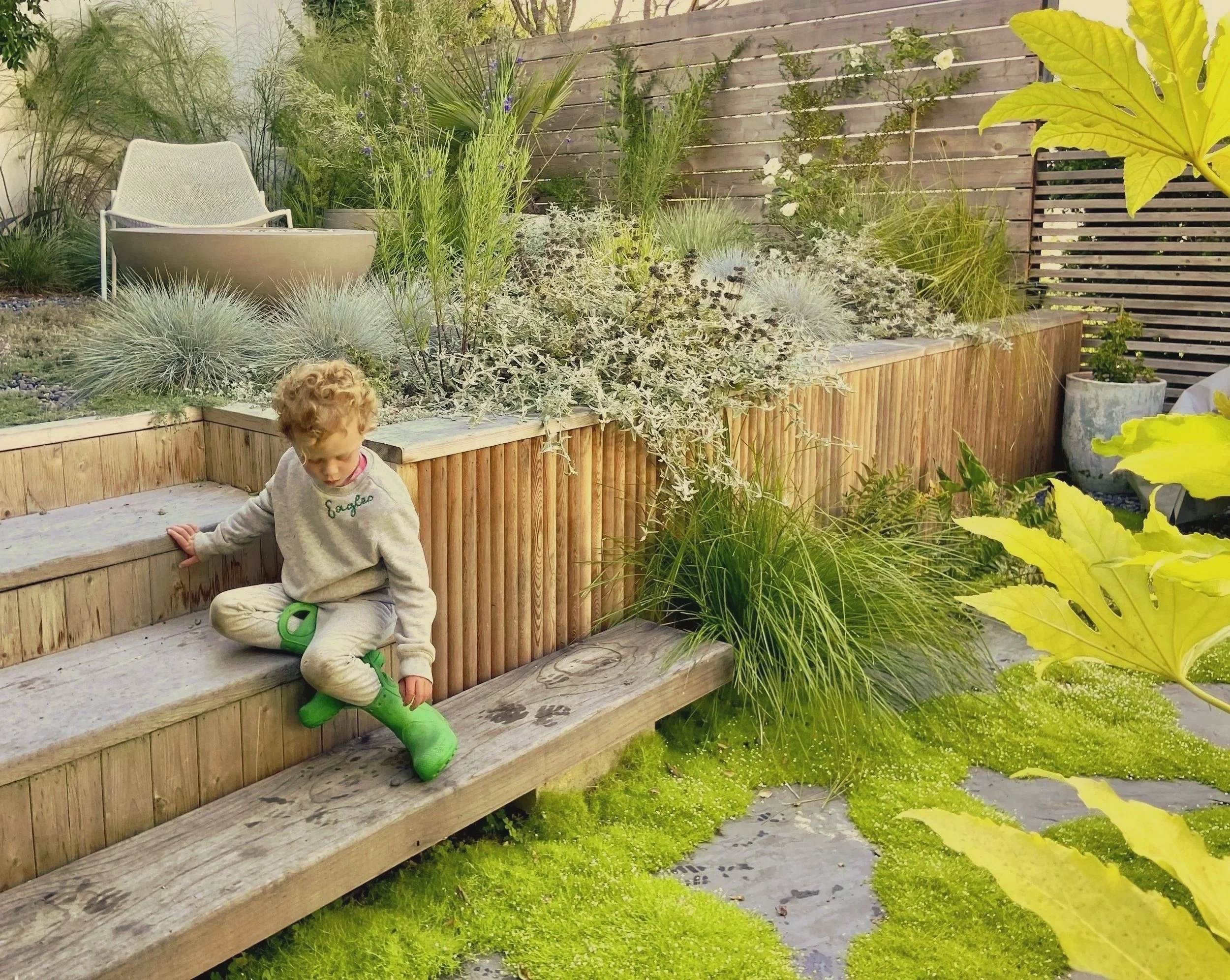 A young child with curly hair sitting on outdoor wooden steps surrounded by lush green plants and shrubs in a landscaped backyard garden.