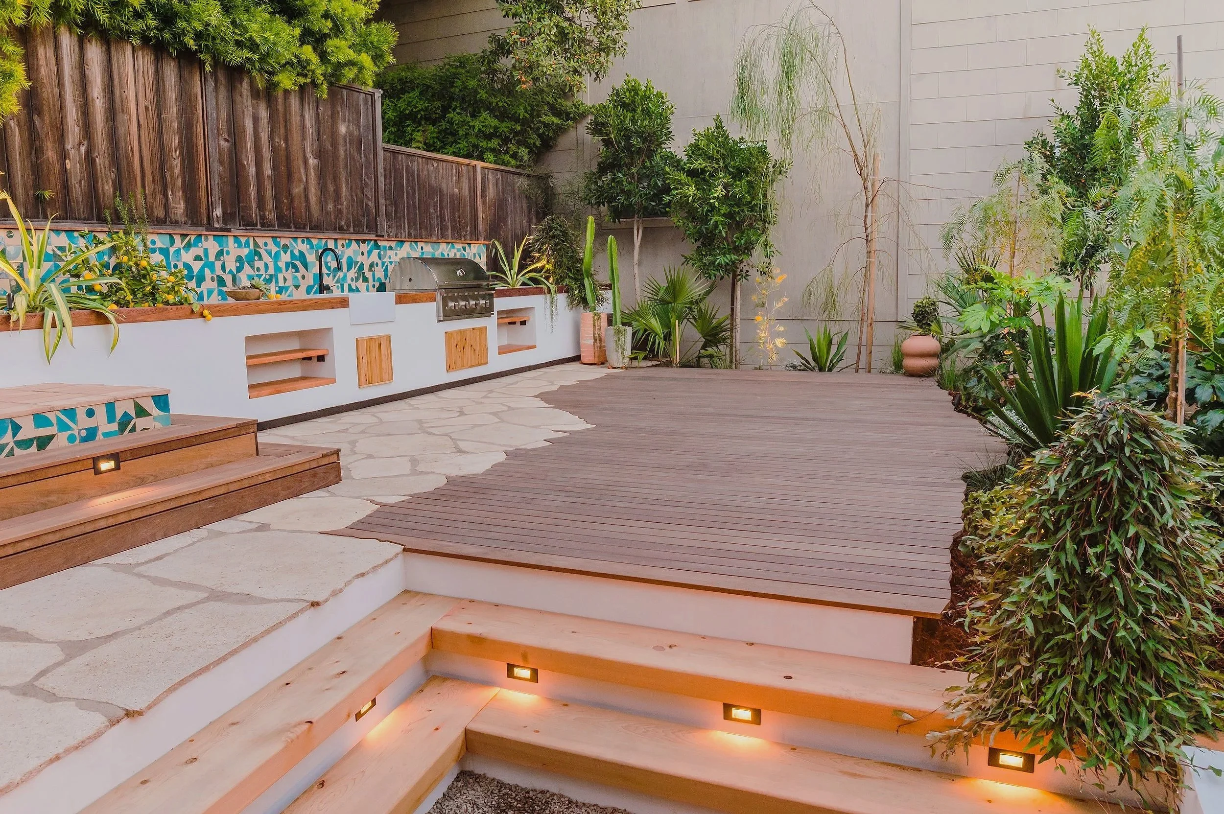 Backyard with wooden deck, stone patio, built-in outdoor kitchen with a grill, and lush green plants and trees along the fence and wall.