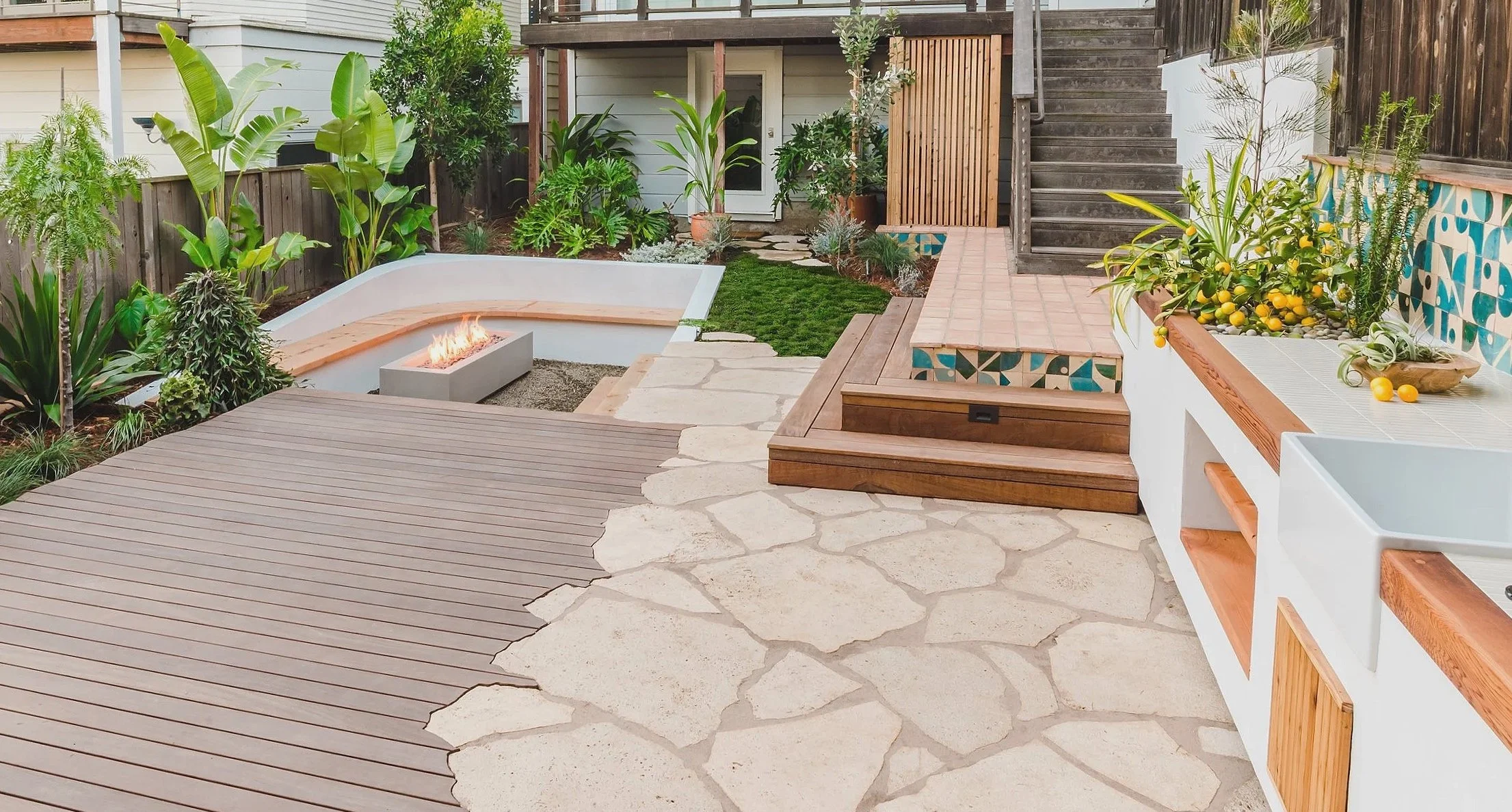 A modern backyard patio with wooden decks, stone walkways, lush plants, a fire pit, and decorative ceramic tiles.