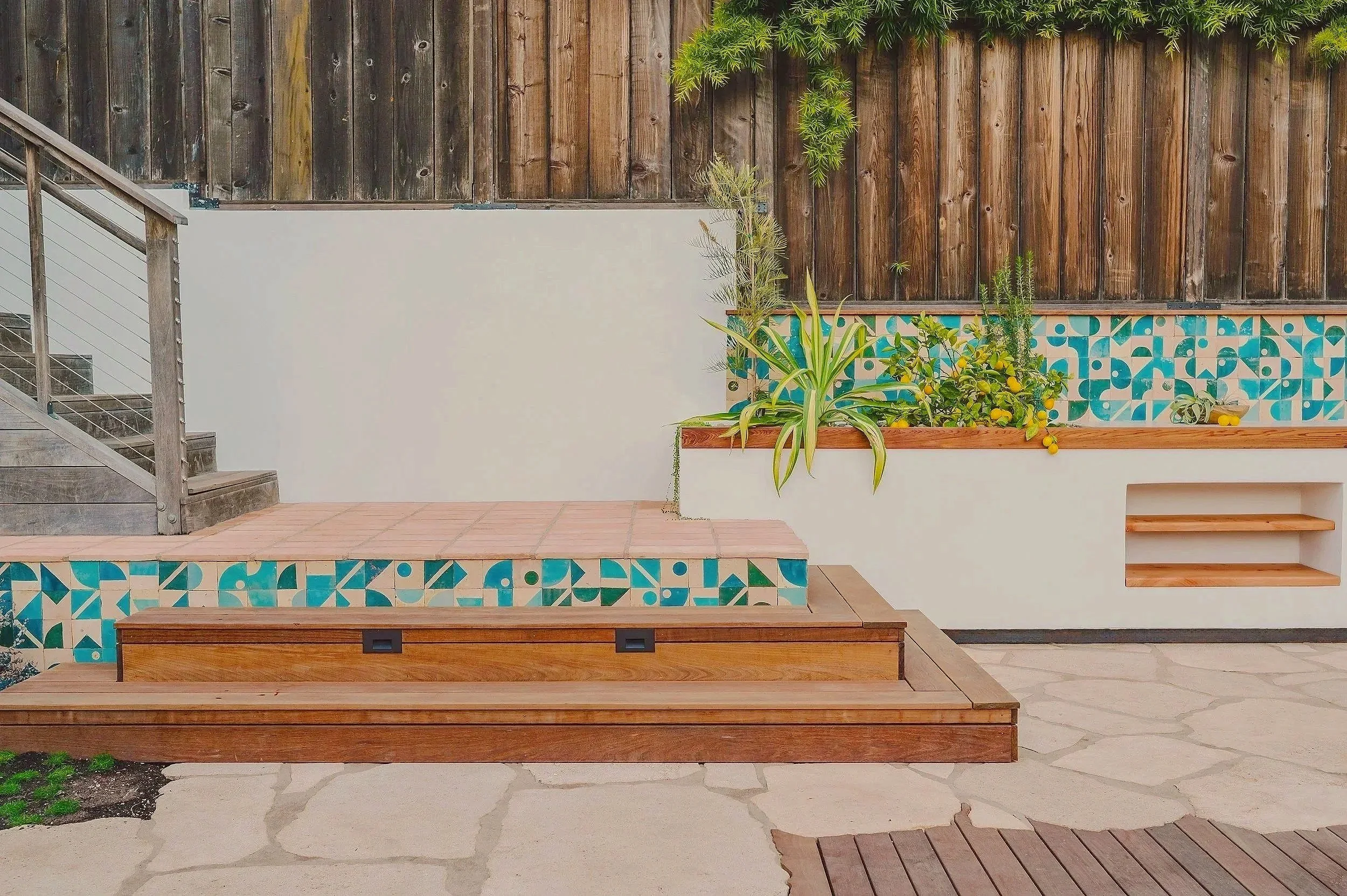 Backyard patio with wooden stairs, decorated with blue and white patterned tiles, green plants, and a wooden fence.