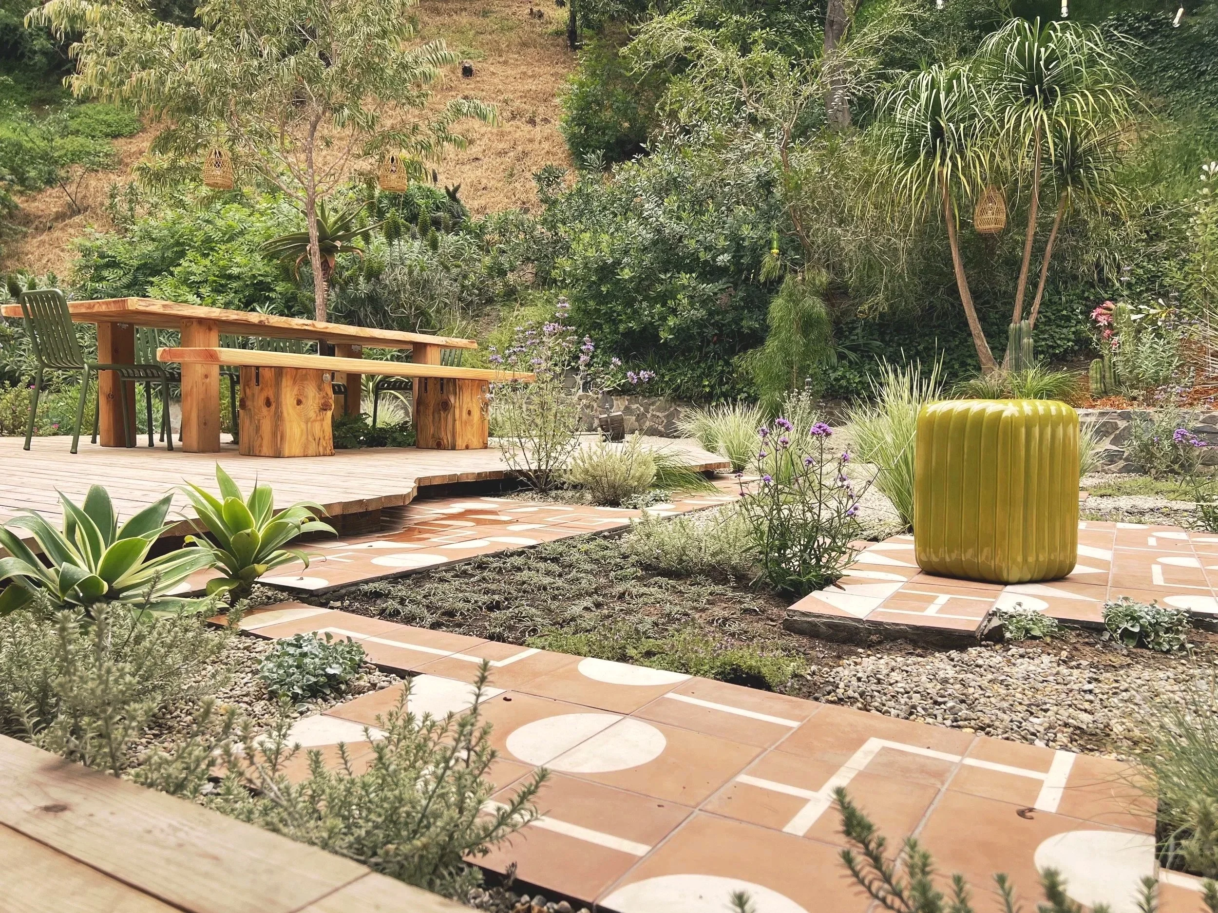 A garden patio featuring a wooden table and benches, a large yellow ceramic stool, greenery with various plants, trees, and decorative hanging lanterns, with a hillside in the background.
