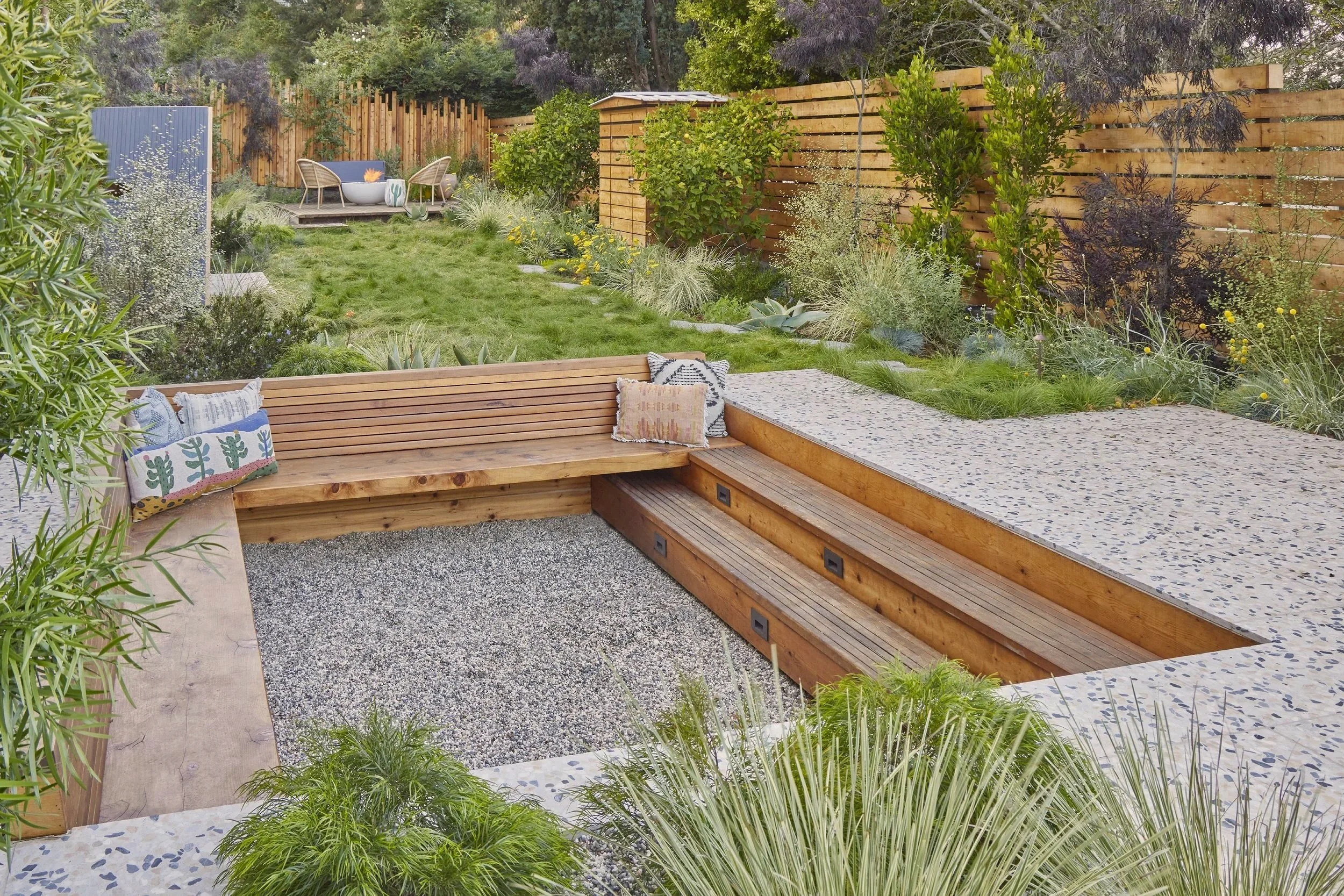 A landscaped backyard garden with a central seating area and stepping stones leading to a grassy area with trees and plants. The backyard features wooden fencing and outdoor furniture.