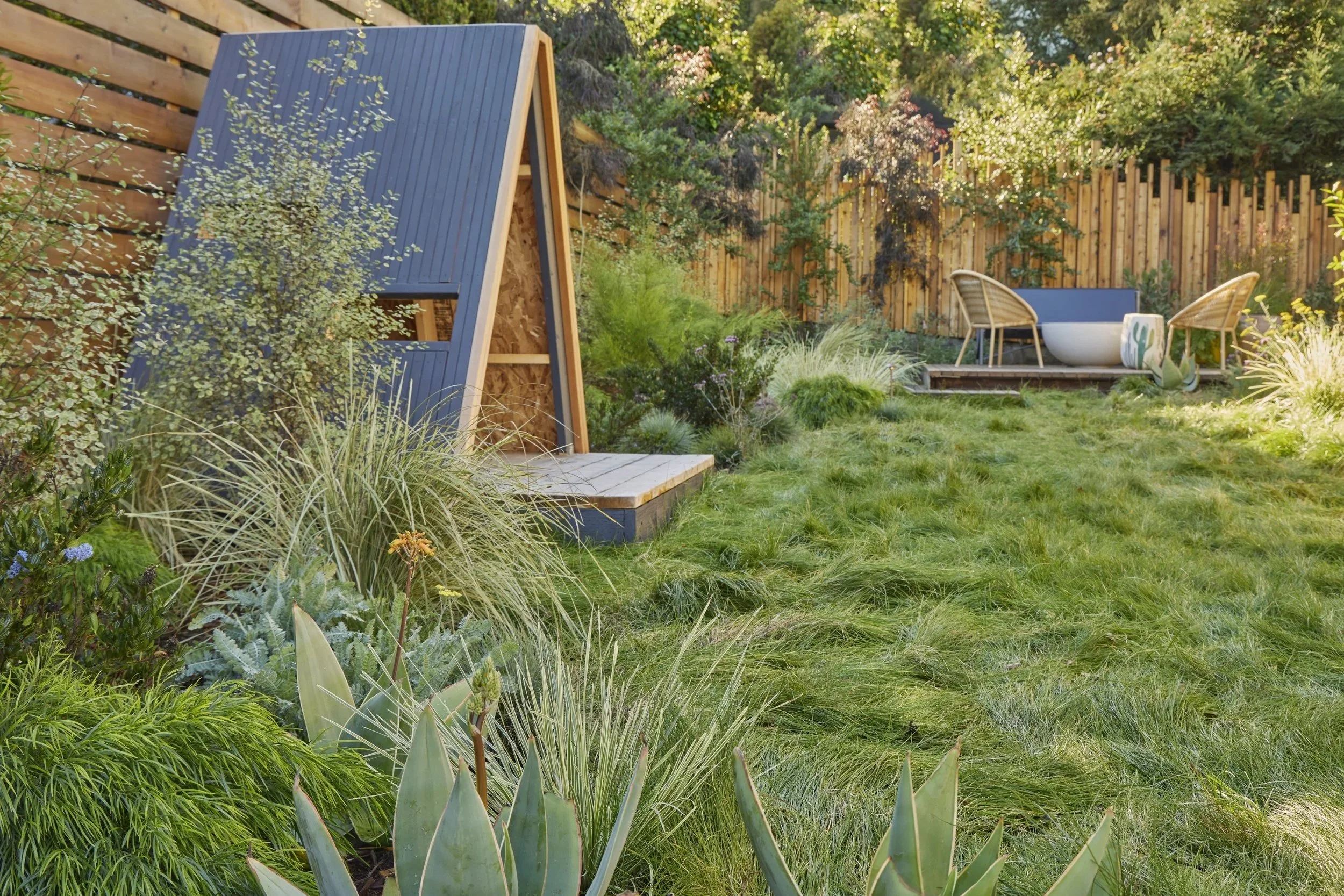 A backyard with a small wooden shed, lush green grass, various plants, flowering bushes, a wooden deck with two chairs, a bathtub, and decorative pots enclosed by a wooden fence.